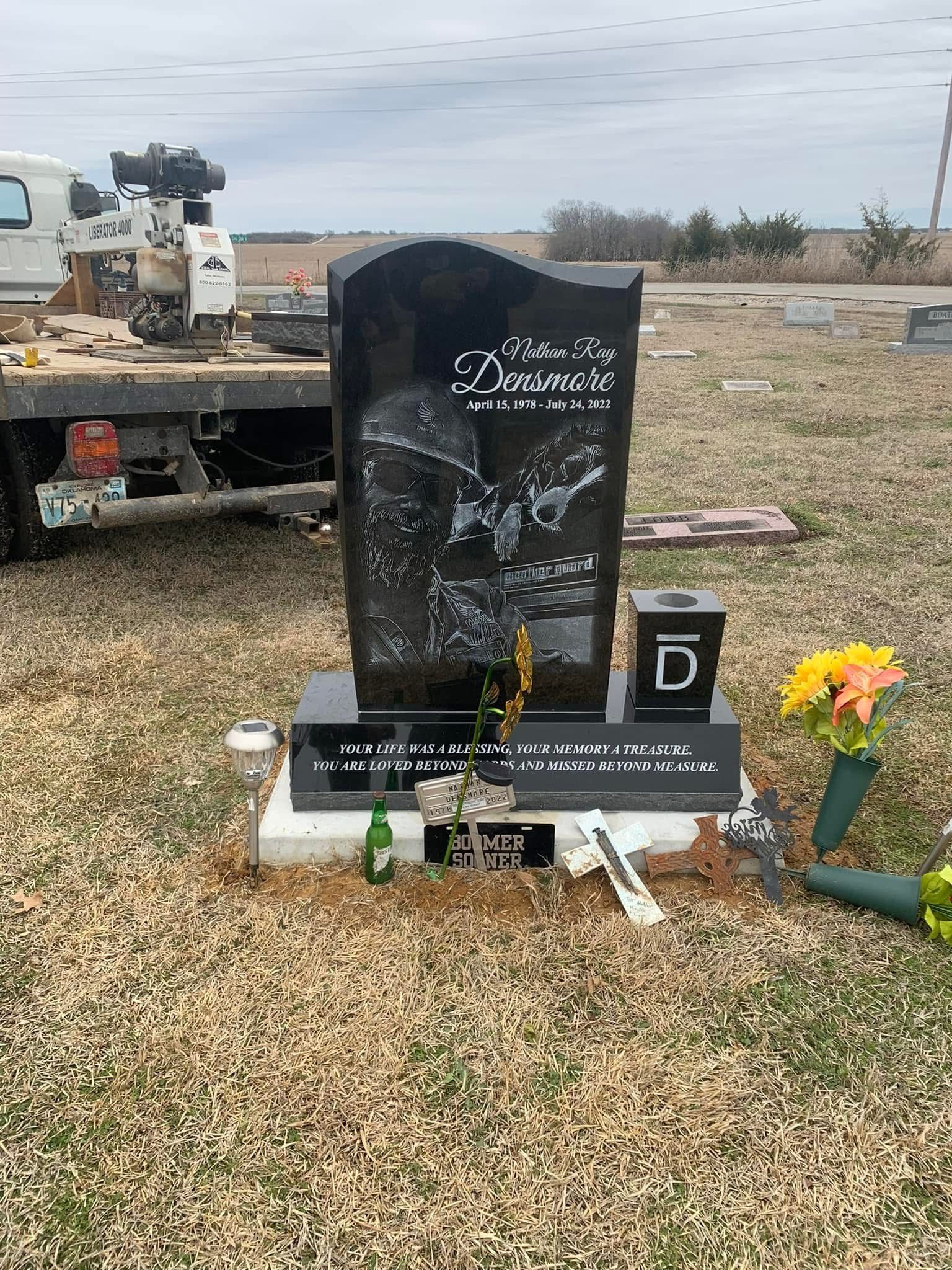 Black headstone with flowers in a cemetery, beside a truck and a small memorial marker.