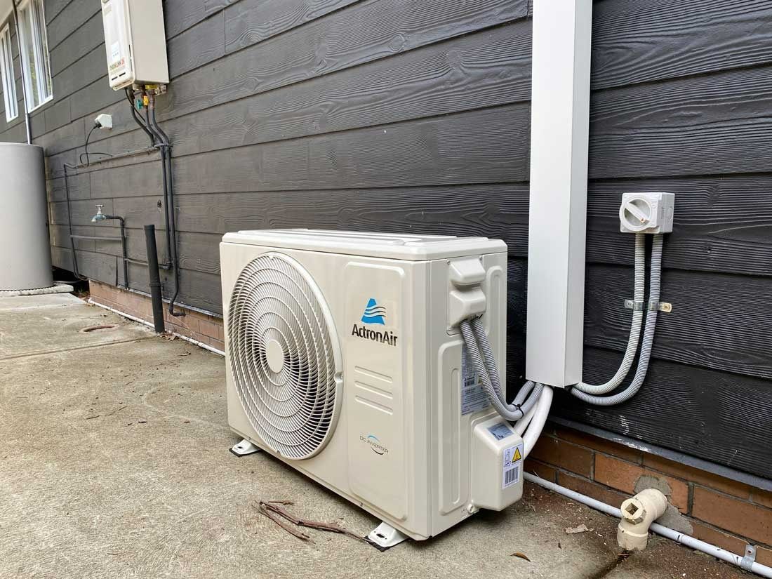 A White Outdoor AC Unit Sits Against a Black Wooden Clad Wall — JF Air Conditioning Pty Ltd in Edgeworth, NSW