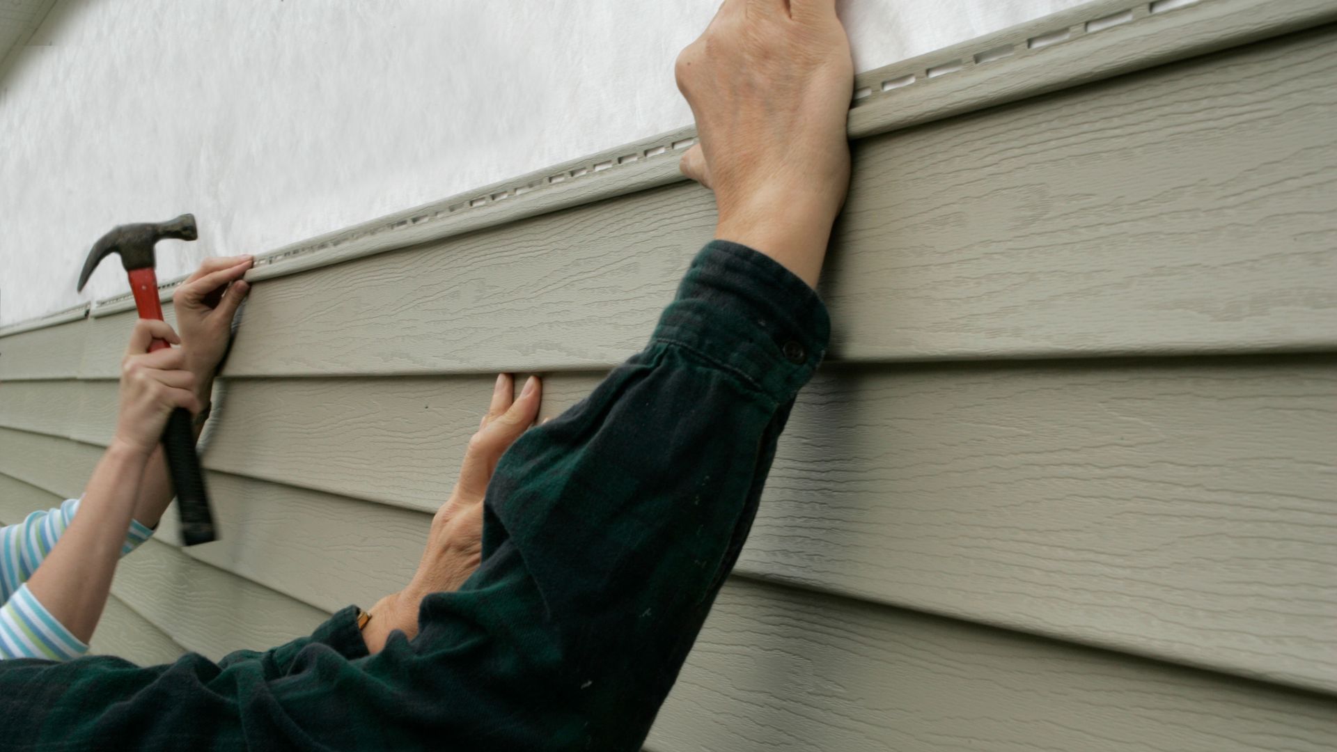 Two people are installing siding on a house with hammers.