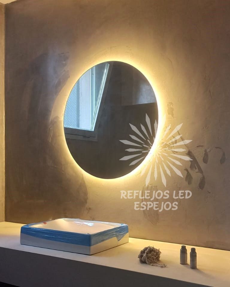 Espejo redondo retroiluminado en la pared con el texto “REFLEJOS LED ESPEJOS” y objetos de exhibición en un estante.