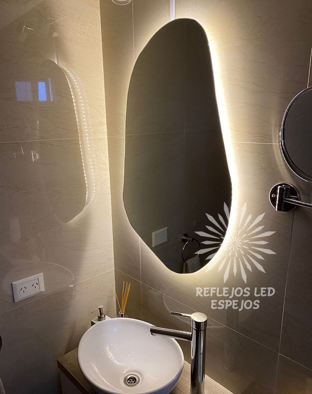 Lavabo moderno con espejo ovalado y pared retroiluminada, lavabo blanco, grifo cromado y texto en español en la pared.