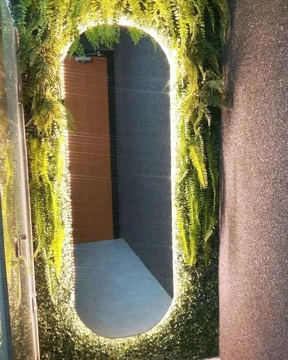 Espejo ovalado enmarcado con follaje verde junto a una pared texturizada, que refleja una puerta y un suelo de baldosas.