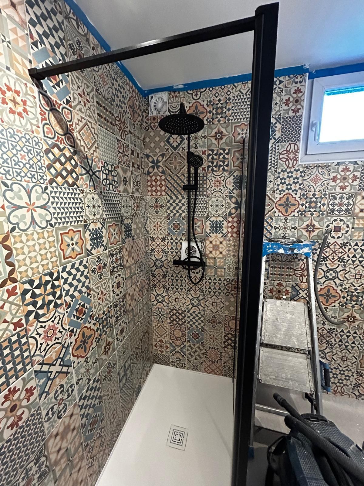 Cabine de douche carrelée avec robinetterie noire, receveur blanc et petite fenêtre.