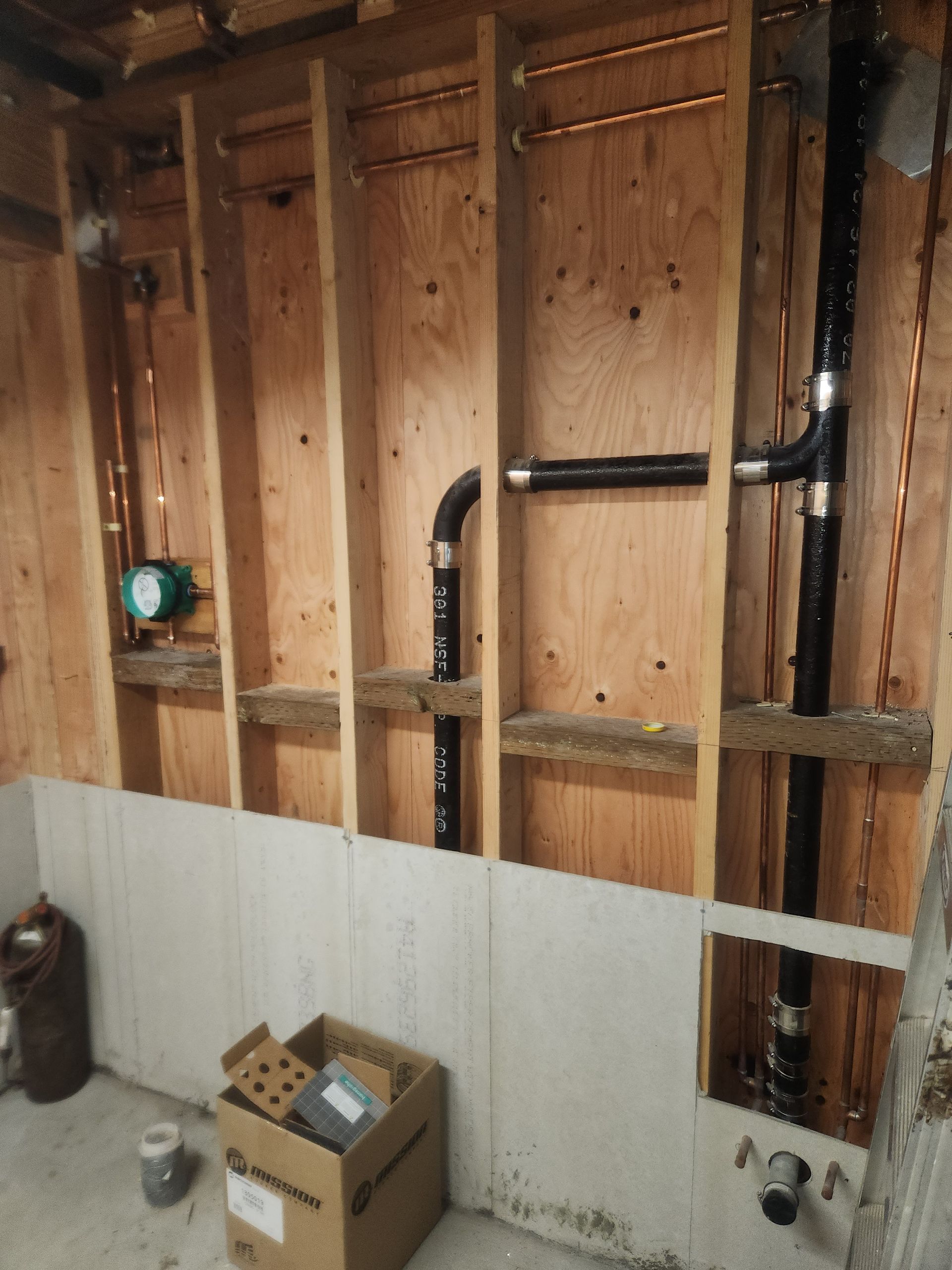 Unfinished basement wall with exposed studs, pipes, wiring, and a few boxes on the floor.