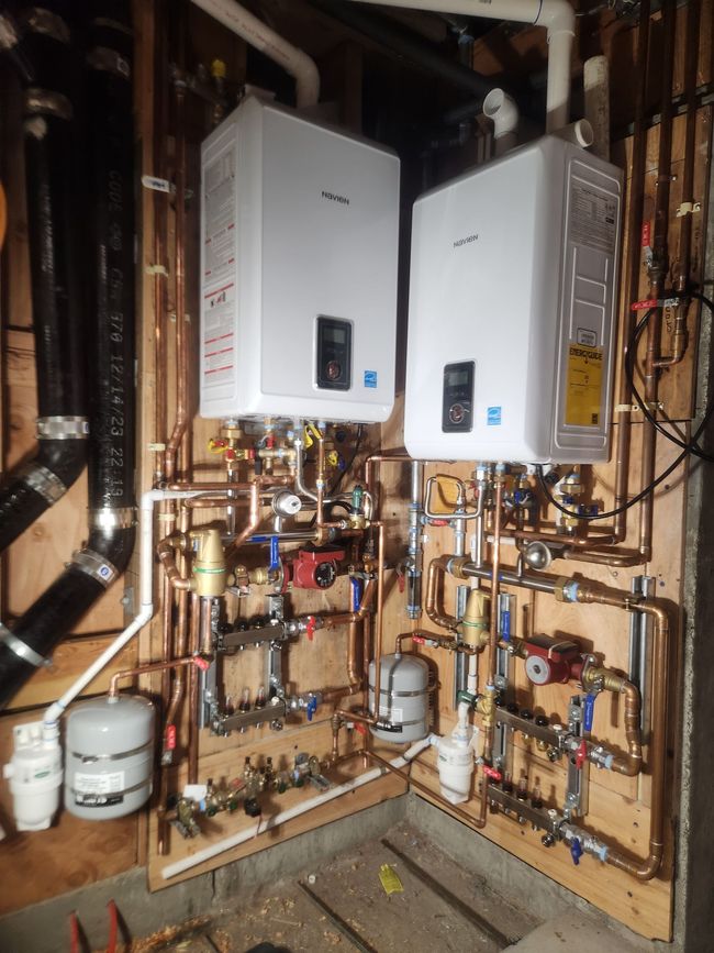 Two white wall-mounted boilers and exposed copper piping in a utility room basement