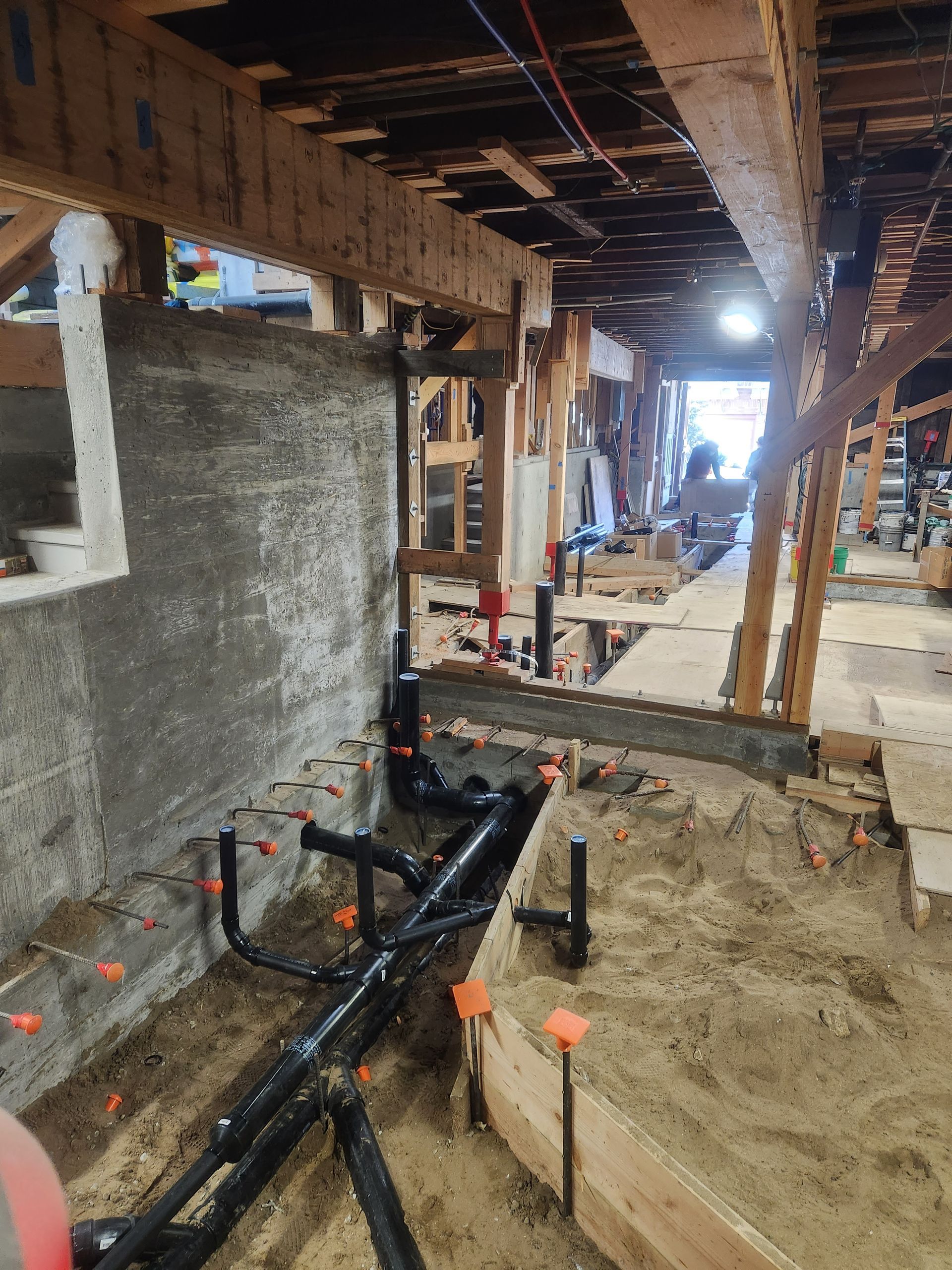 Construction site interior with exposed beams, plumbing, and a sandy floor under renovation