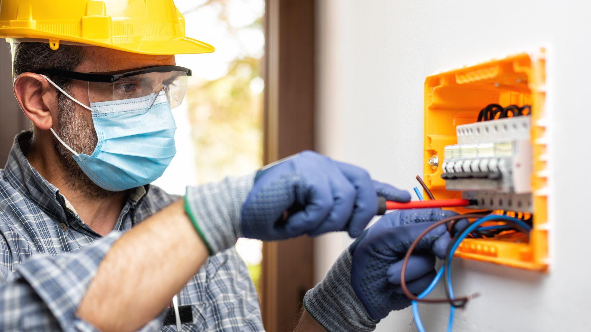 An electrician wearing a mask and gloves is working on an electrical box.