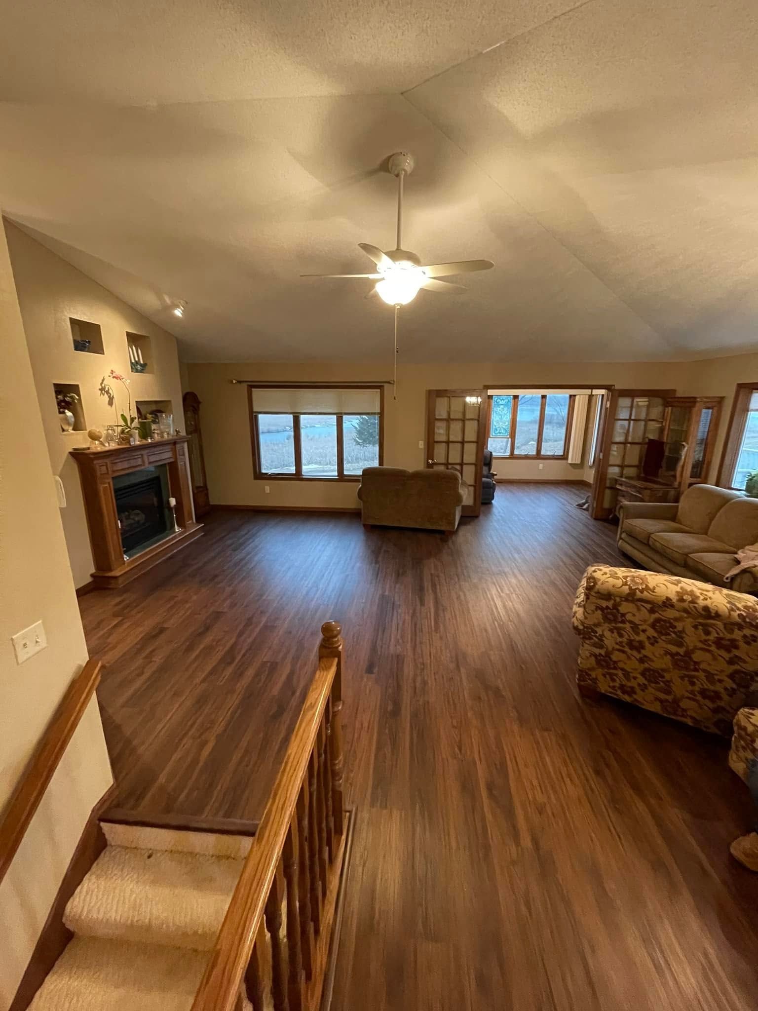 Spacious living room with wood floors, fireplace, ceiling fan, and large windows.
