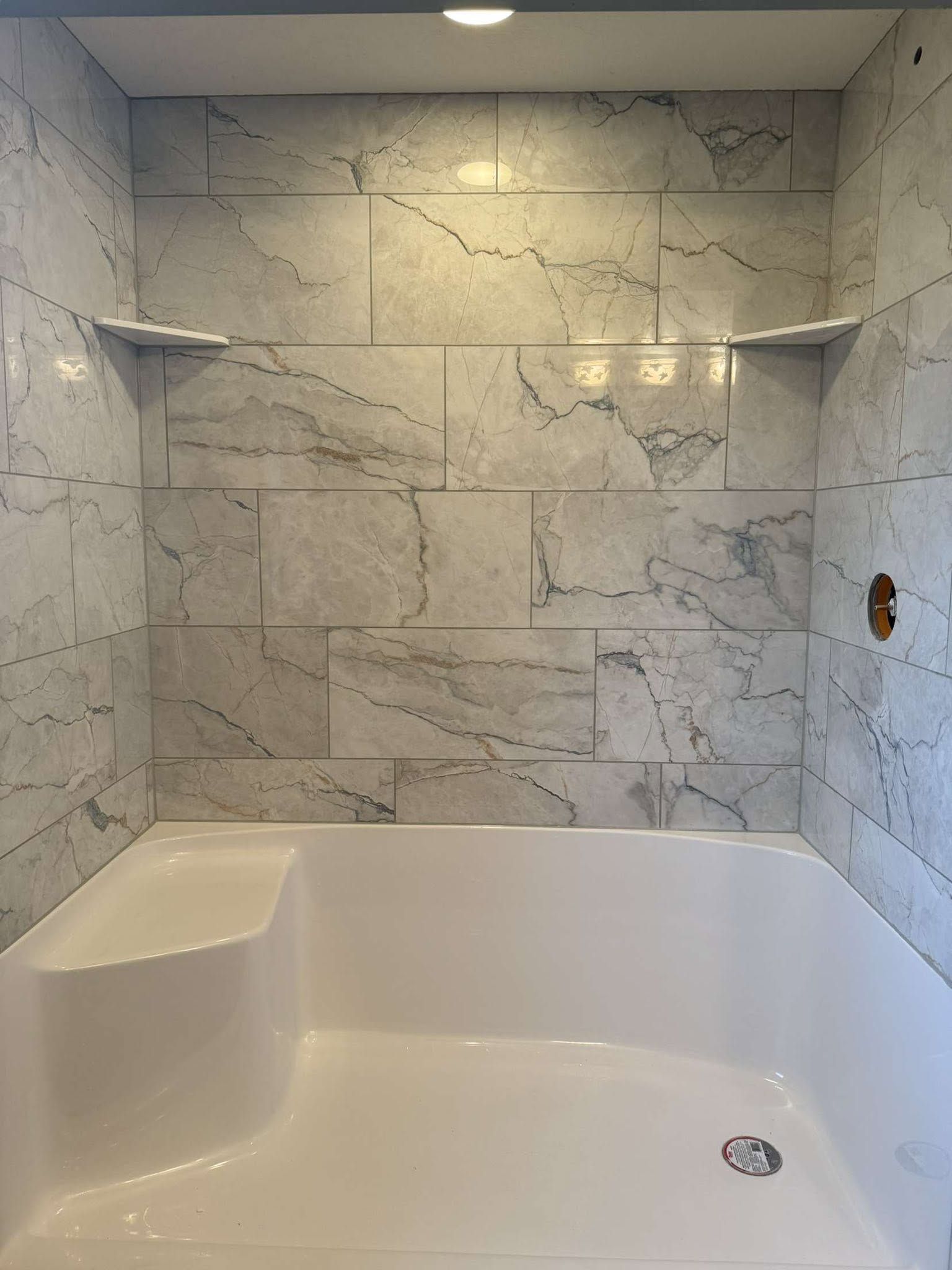 Modern white marble-tiled bathroom with a white tub and built-in corner shelf under recessed lighting