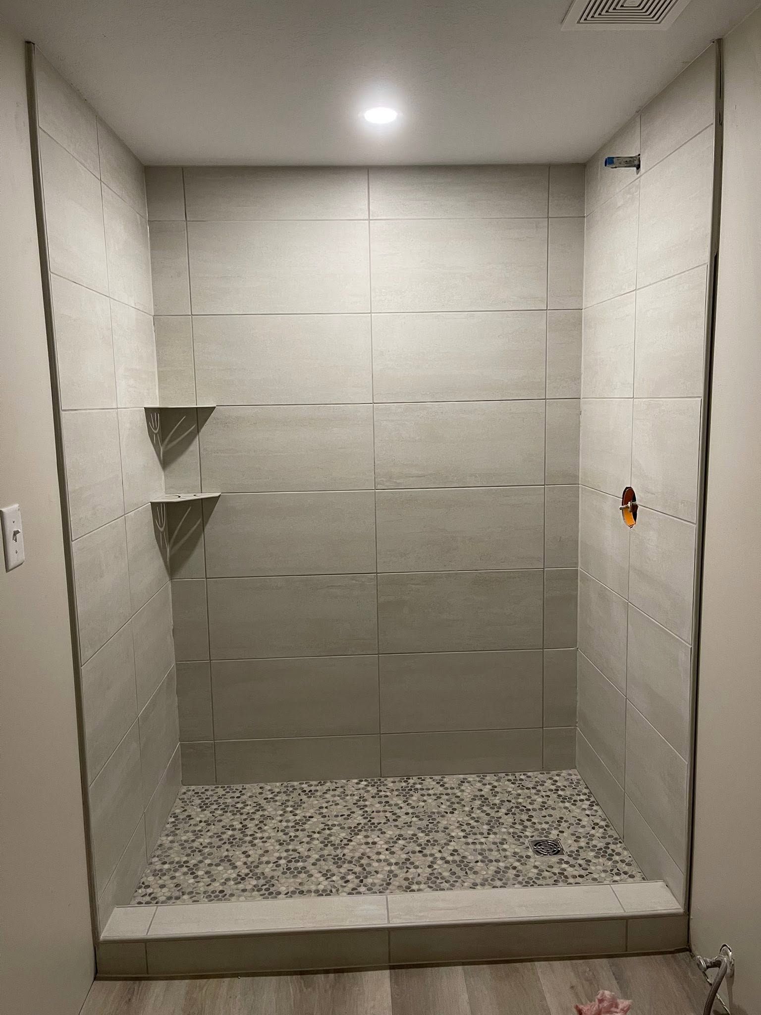 Small tiled shower stall with a pebble floor and two corner shelves.