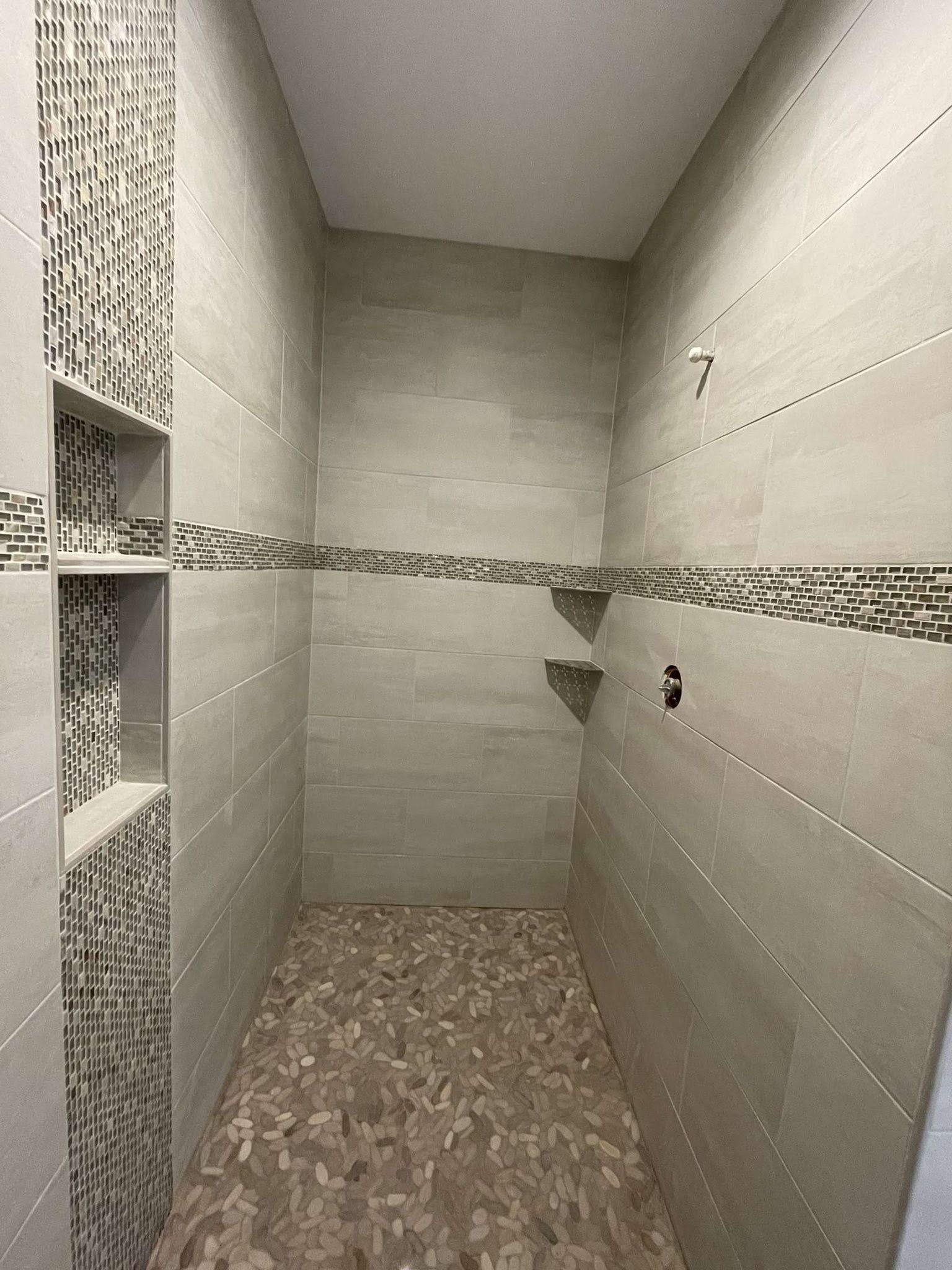 Narrow tiled shower stall with gray walls, mosaic accent strip, and built-in shelf niches.