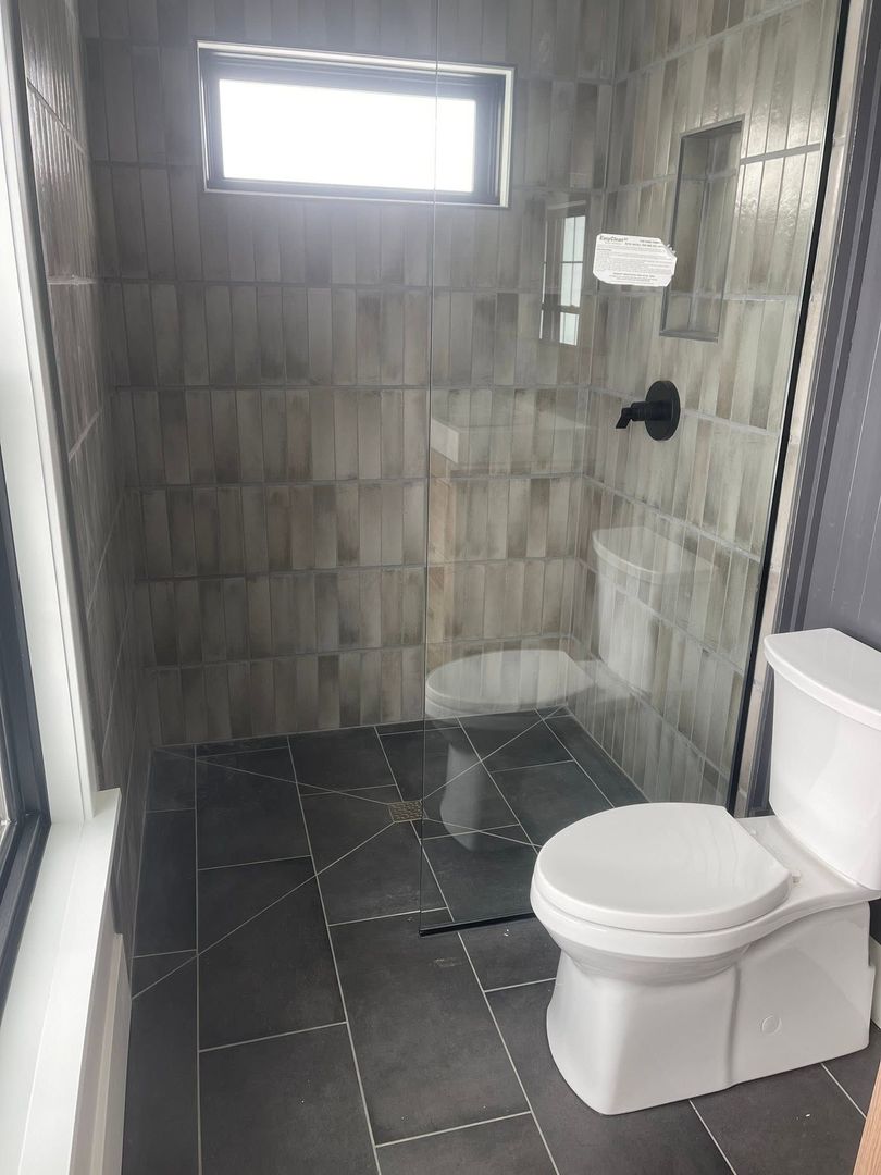 Small tiled bathroom with a toilet, glass shower, and narrow frosted window.