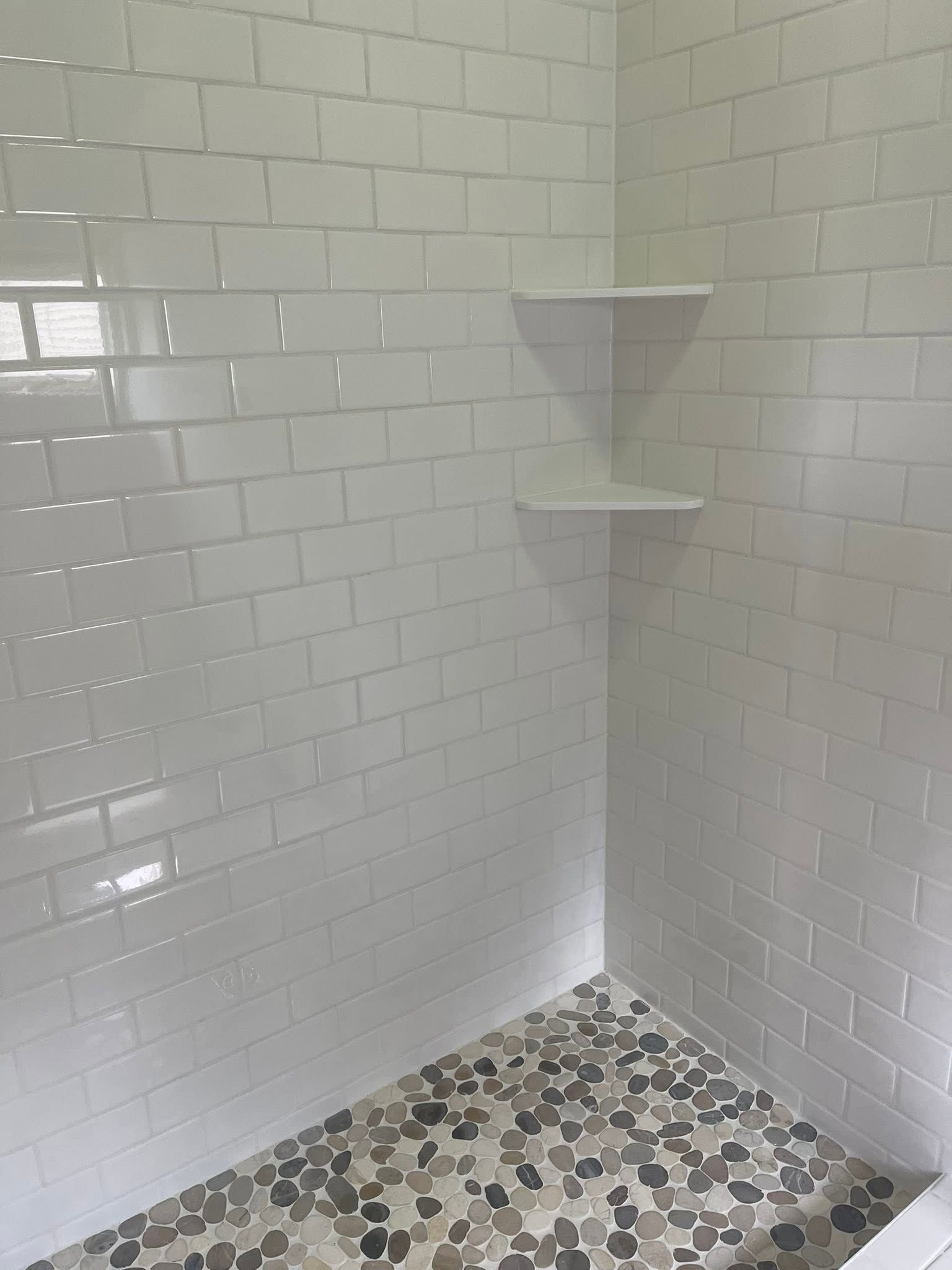 White tiled shower corner with two small shelves and a pebble-pattern floor.