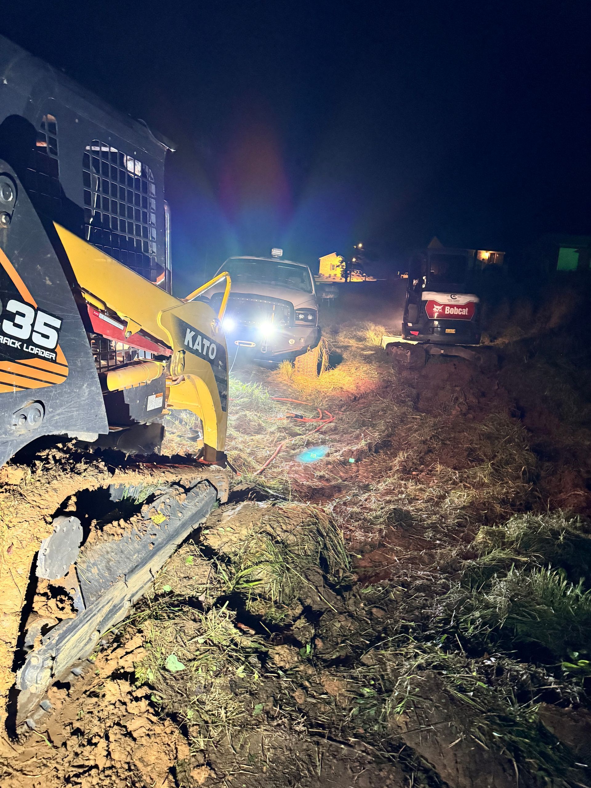 Bobcat excavator and vehicles with headlights on at night, near a building. Construction scene.