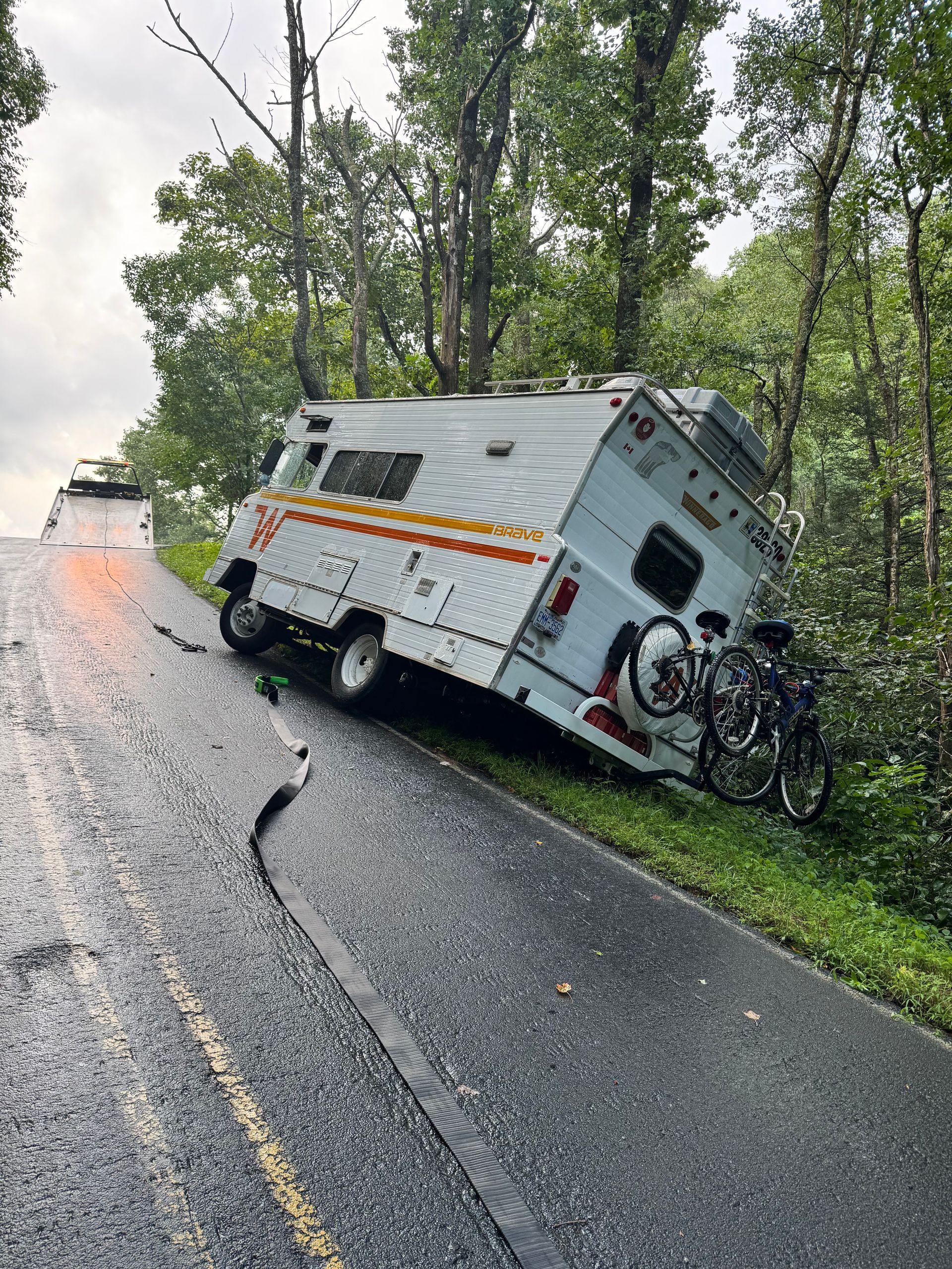 White RV tilted on side of wet road, tires off pavement. Bike leaned against side, trees in background.