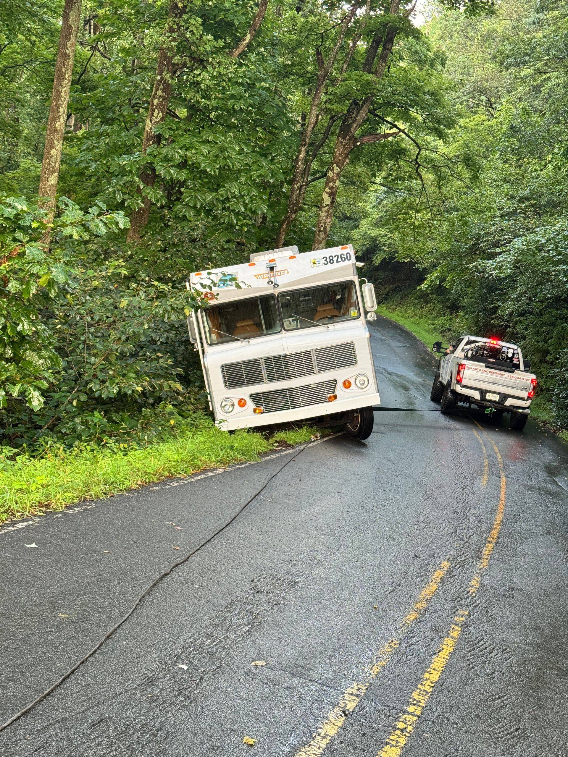 Ice cream truck on a narrow, wet road, partially off the side. A tow truck is present.