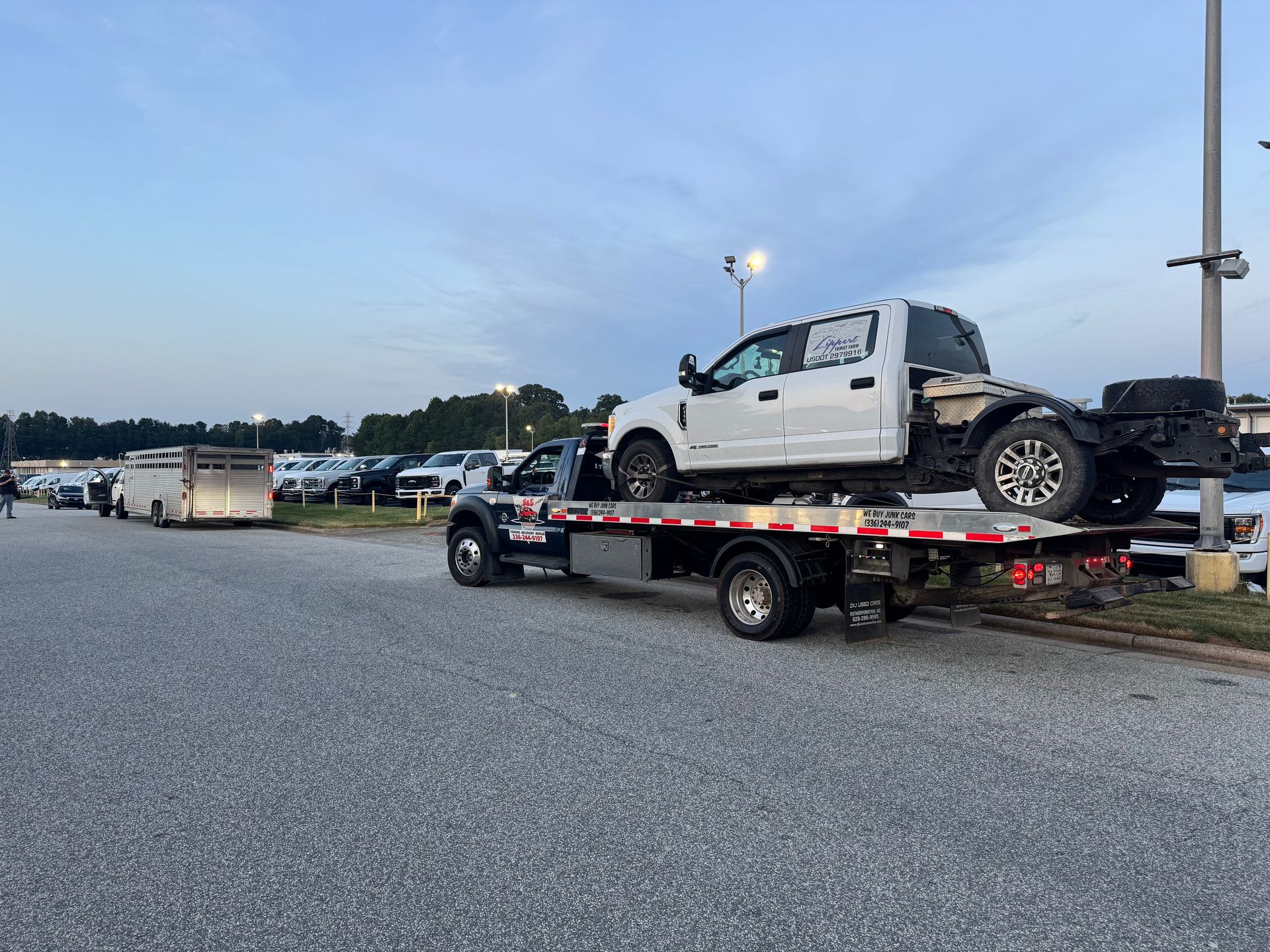 Tow truck carrying a white pickup truck bed and chassis on a paved lot; gray sky.