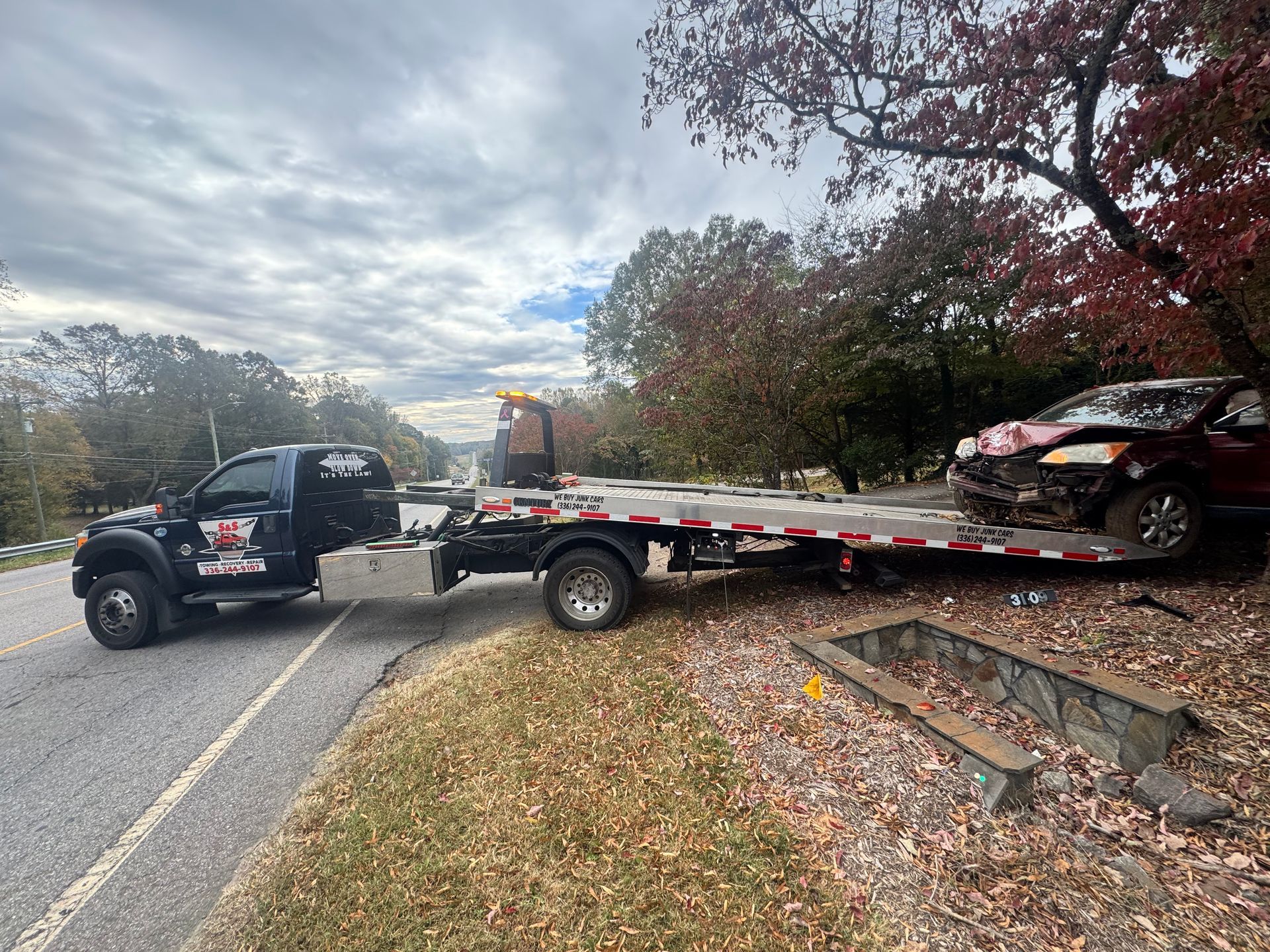 Tow truck loading damaged red car on a road with trees. Overcast sky.