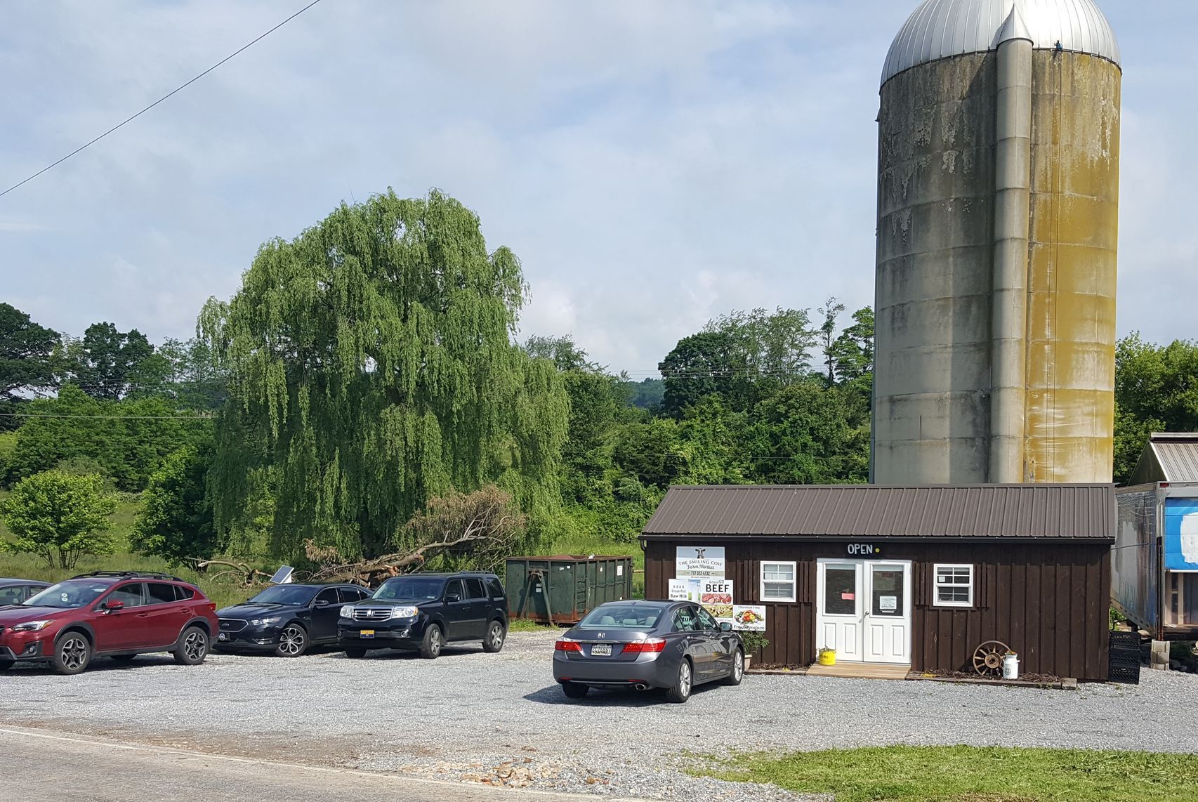 Cars parked in front of a small farm stand with a silo in the background. Lush green trees and a partly cloudy sky.