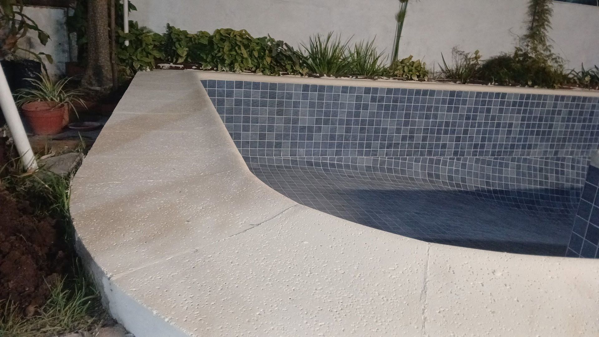 Piscina curva vacía en el patio trasero, revestida con azulejos azules y con un borde de tarima blanca texturizada