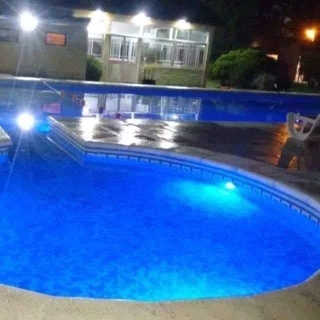 Piscina iluminada en azul por la noche, con un edificio y un patio iluminado al fondo