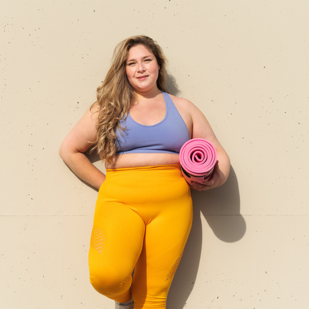 A woman in a blue bra and yellow pants is holding a pink yoga mat.