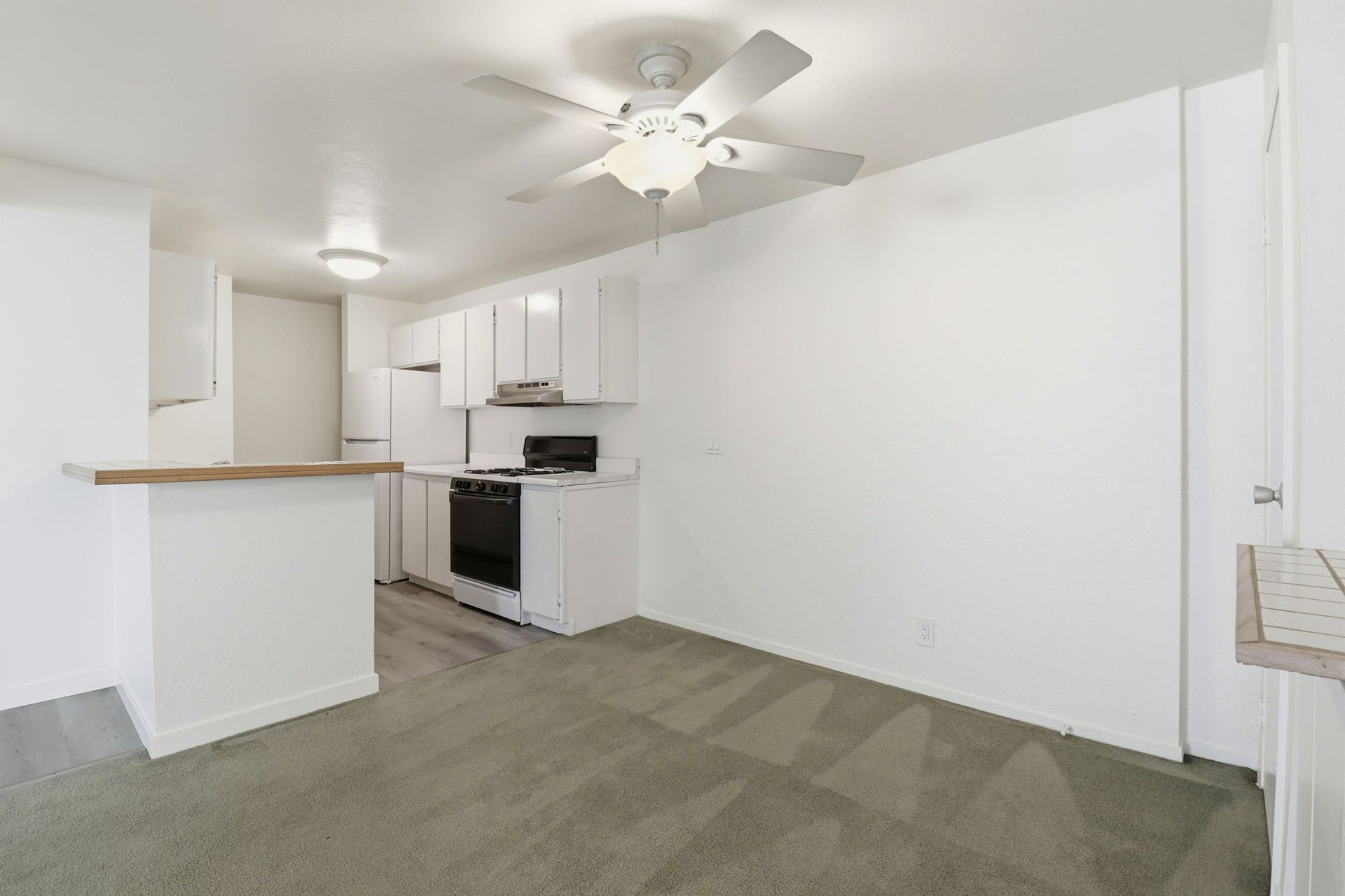 Empty white apartment living room with carpet, ceiling fan, and small kitchen with breakfast bar.