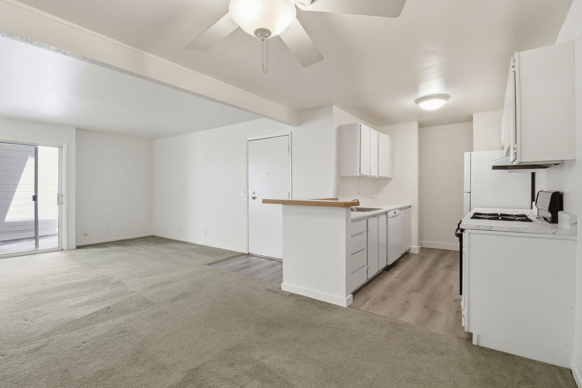 Bright vacant apartment with open kitchen, white cabinets, gray carpet, and ceiling fans