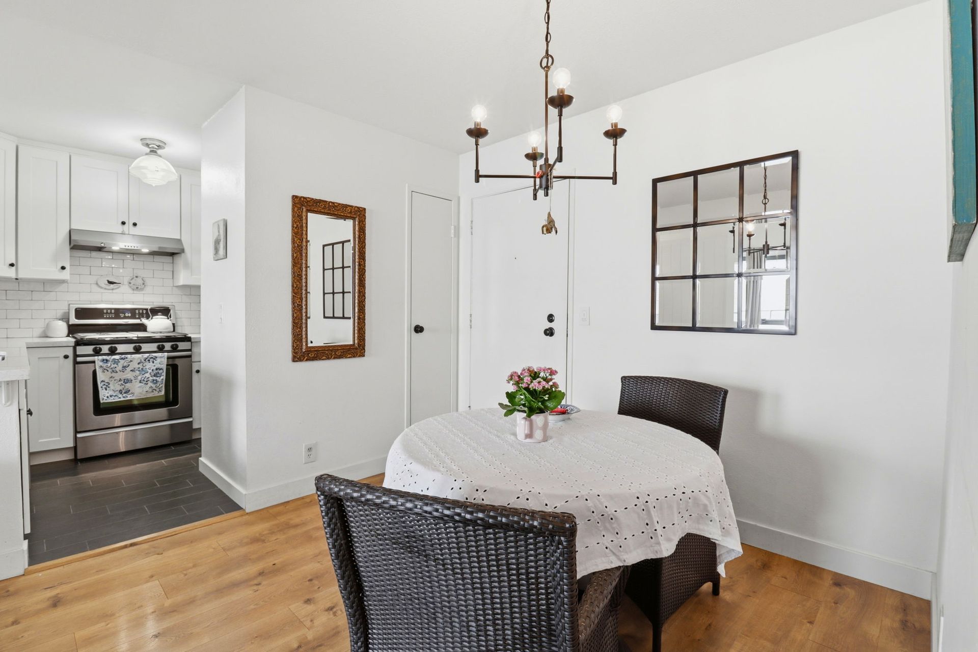 Bright dining room with round table, wicker chairs, chandelier, and a view into a white kitchen.