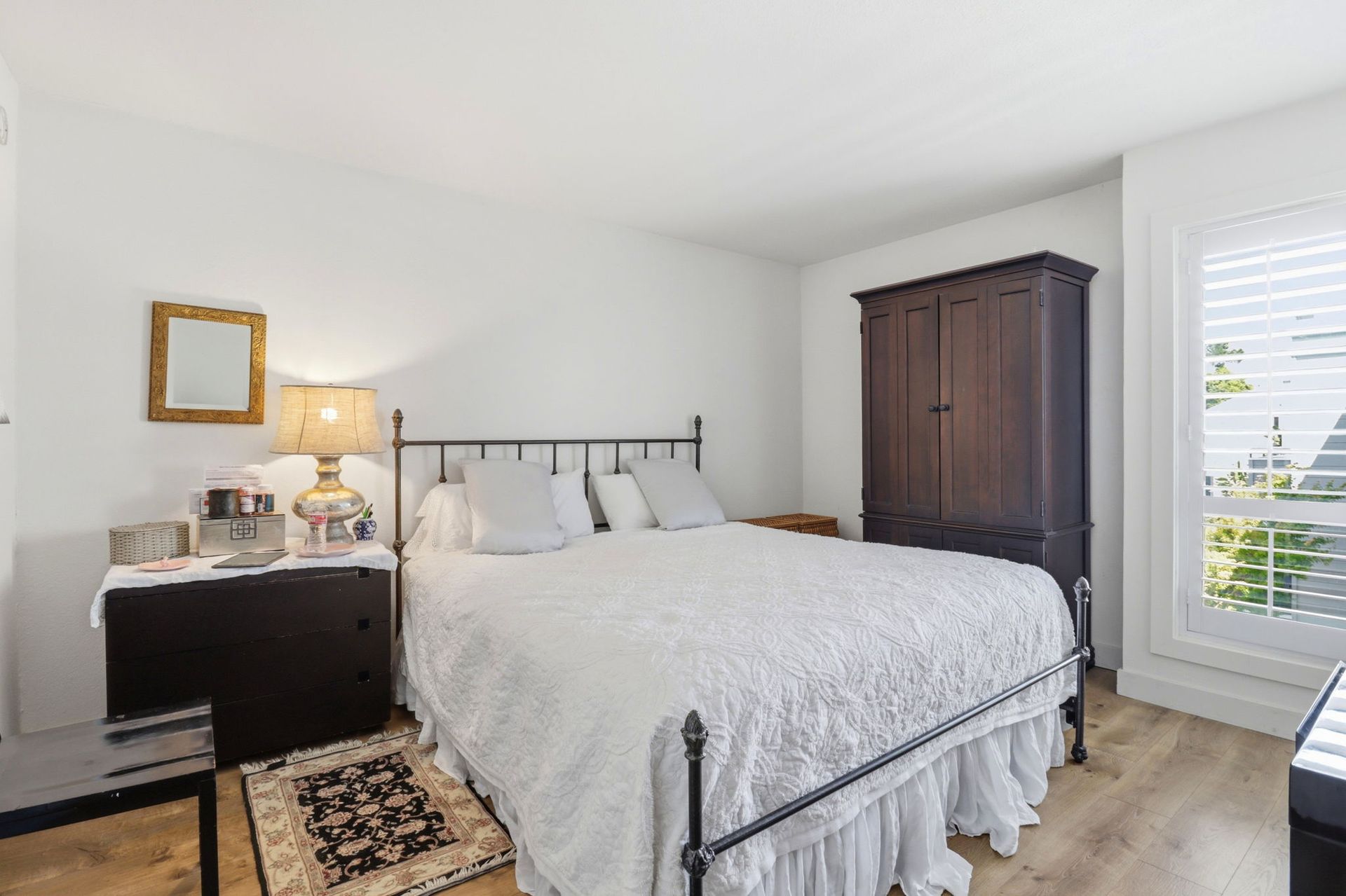 Bright bedroom with white bedding, dark wood dresser and wardrobe, nightstand lamp, and window blinds