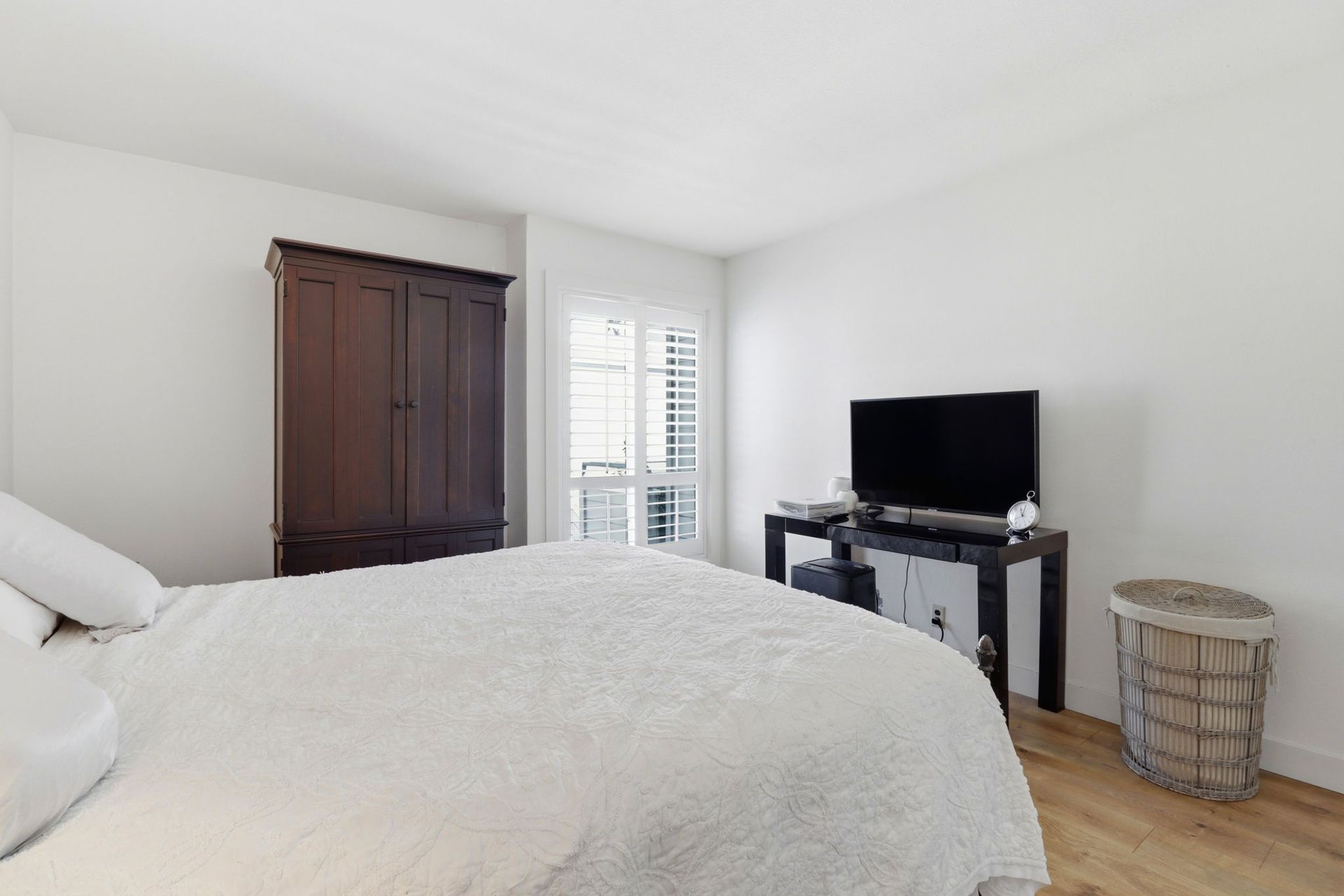 Bright bedroom with a large bed, dark wooden wardrobe, TV on a stand, and wicker hamper by the wall.