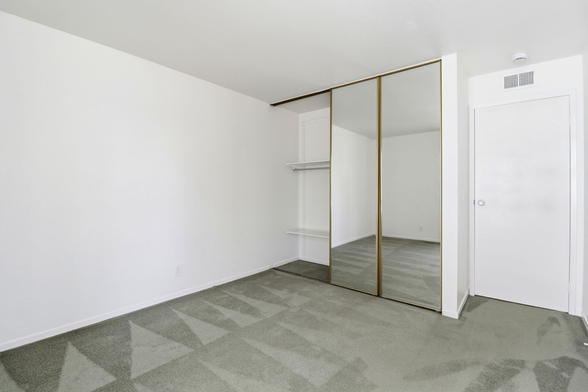 Empty white bedroom with mirrored closet doors, green carpet, and a closed door