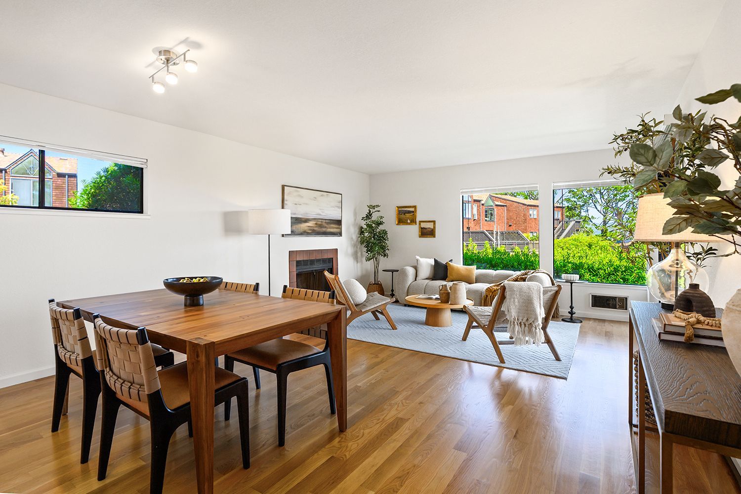 Bright open dining and living room with wooden table, gray sofa, and large windows overlooking greenery