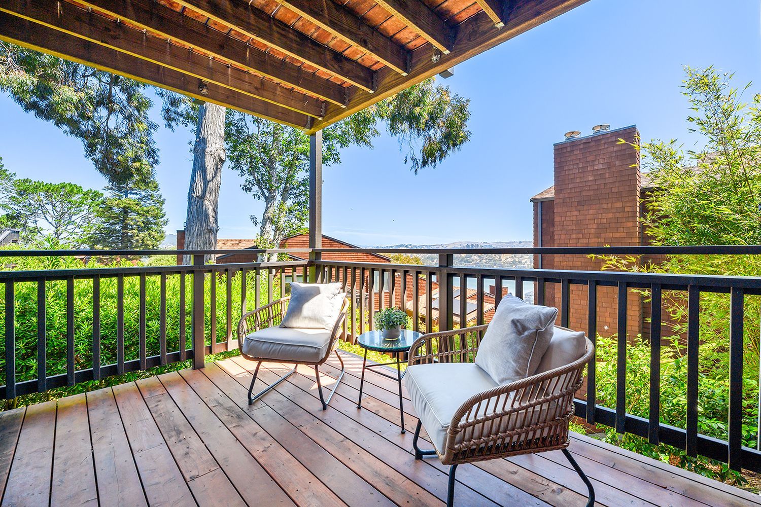 Covered wooden balcony with two lounge chairs, railings, and green trees under a bright sky.