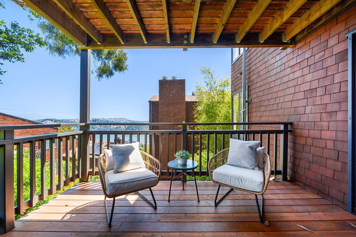 Wooden balcony with two cushioned chairs, a small table, and a city view beyond the railing.