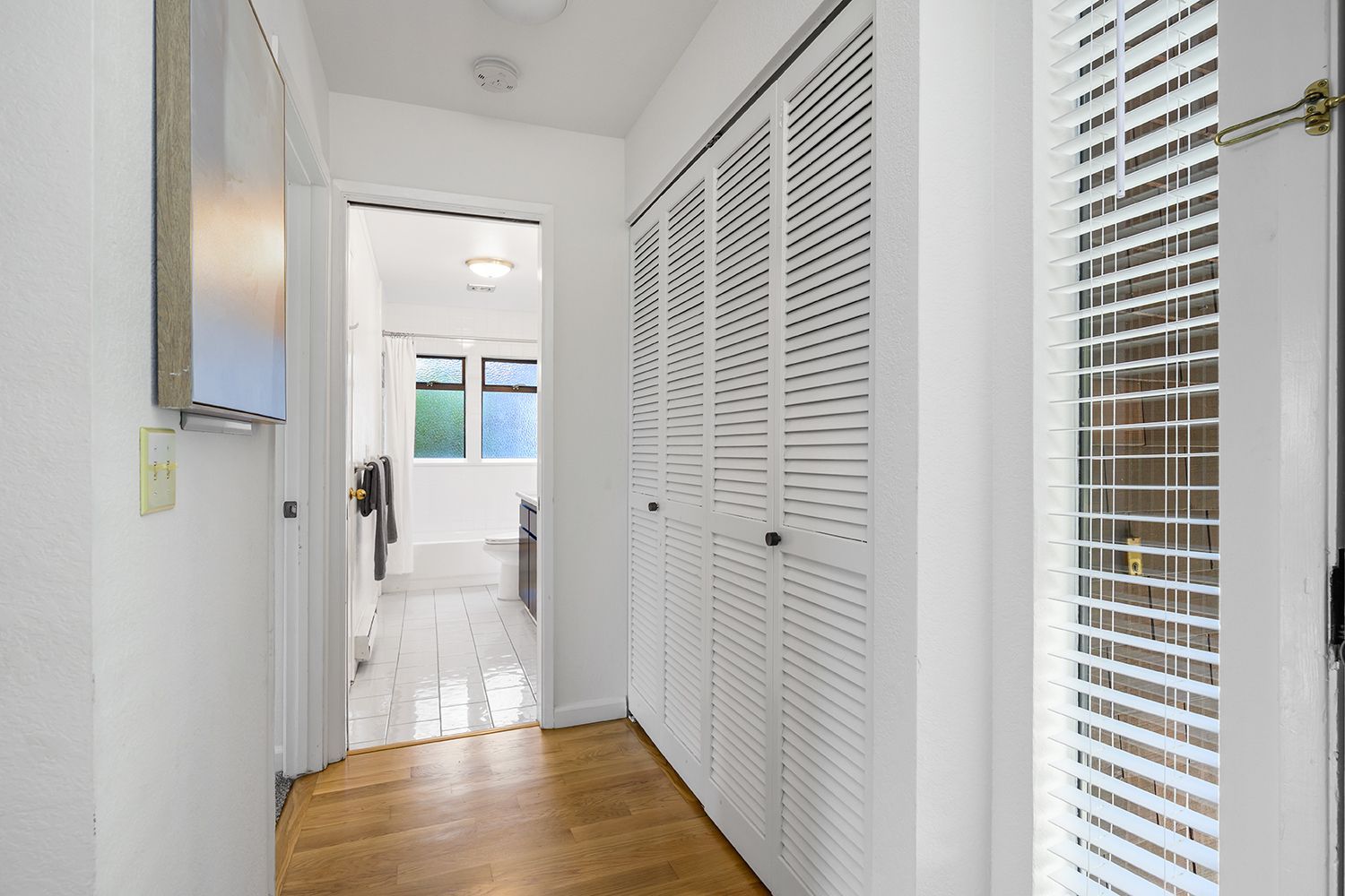 Bright hallway with white louvered closet doors, wood floor, and a view into a sunlit room ahead