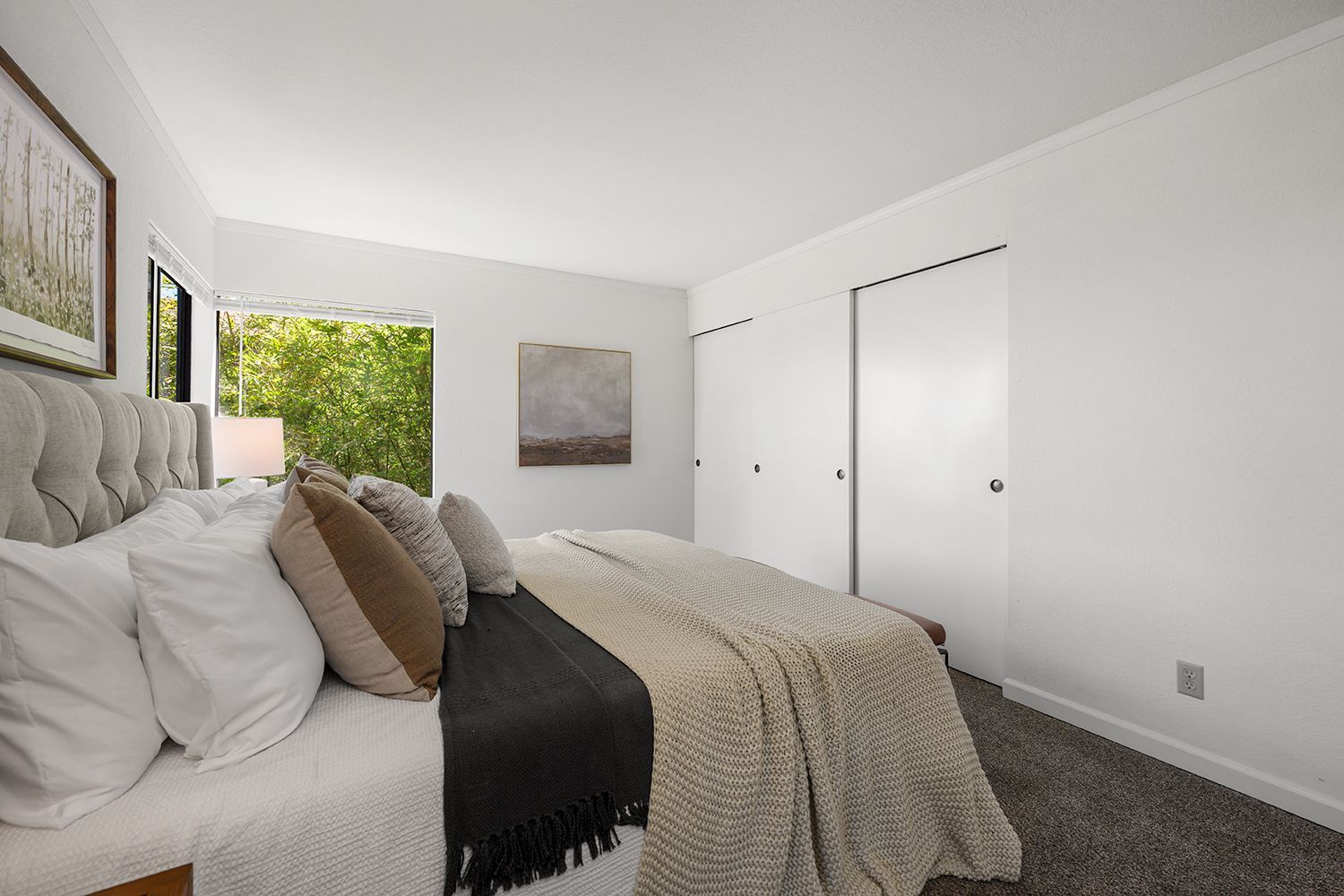 Bright bedroom with a tufted bed, white closet doors, and a window showing greenery.