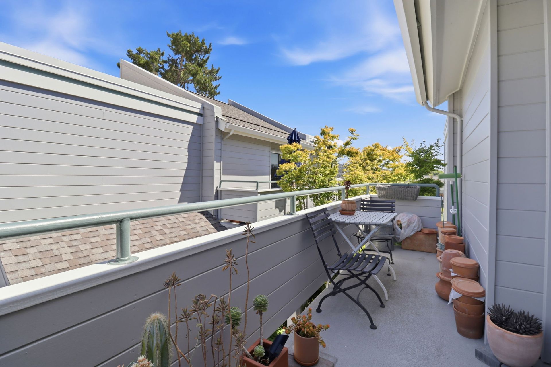 Narrow apartment balcony with metal railing, patio chairs, potted plants, and a view of neighboring buildings.