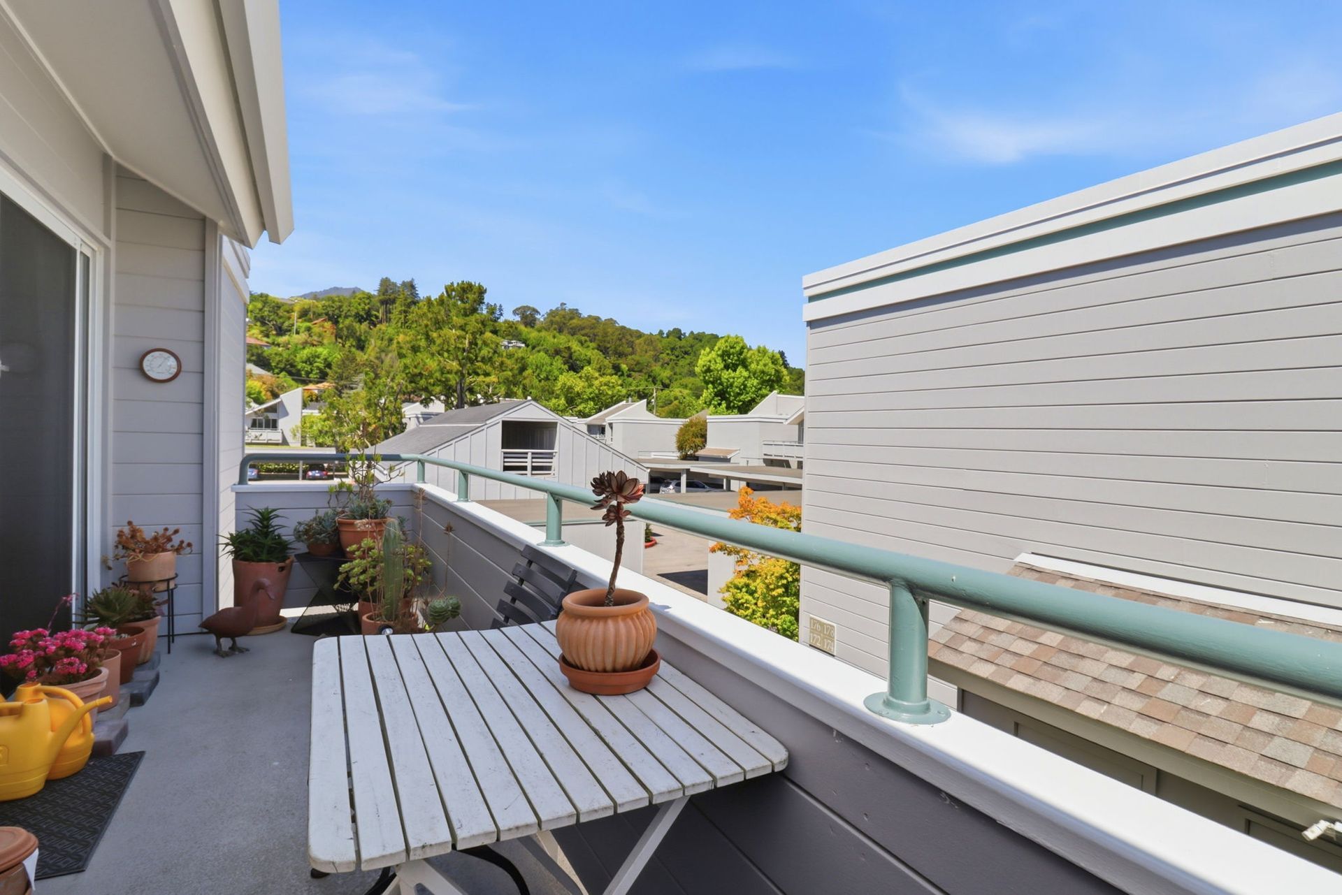 Small balcony with gray table, potted plants, and white railing overlooking neighboring homes and trees.