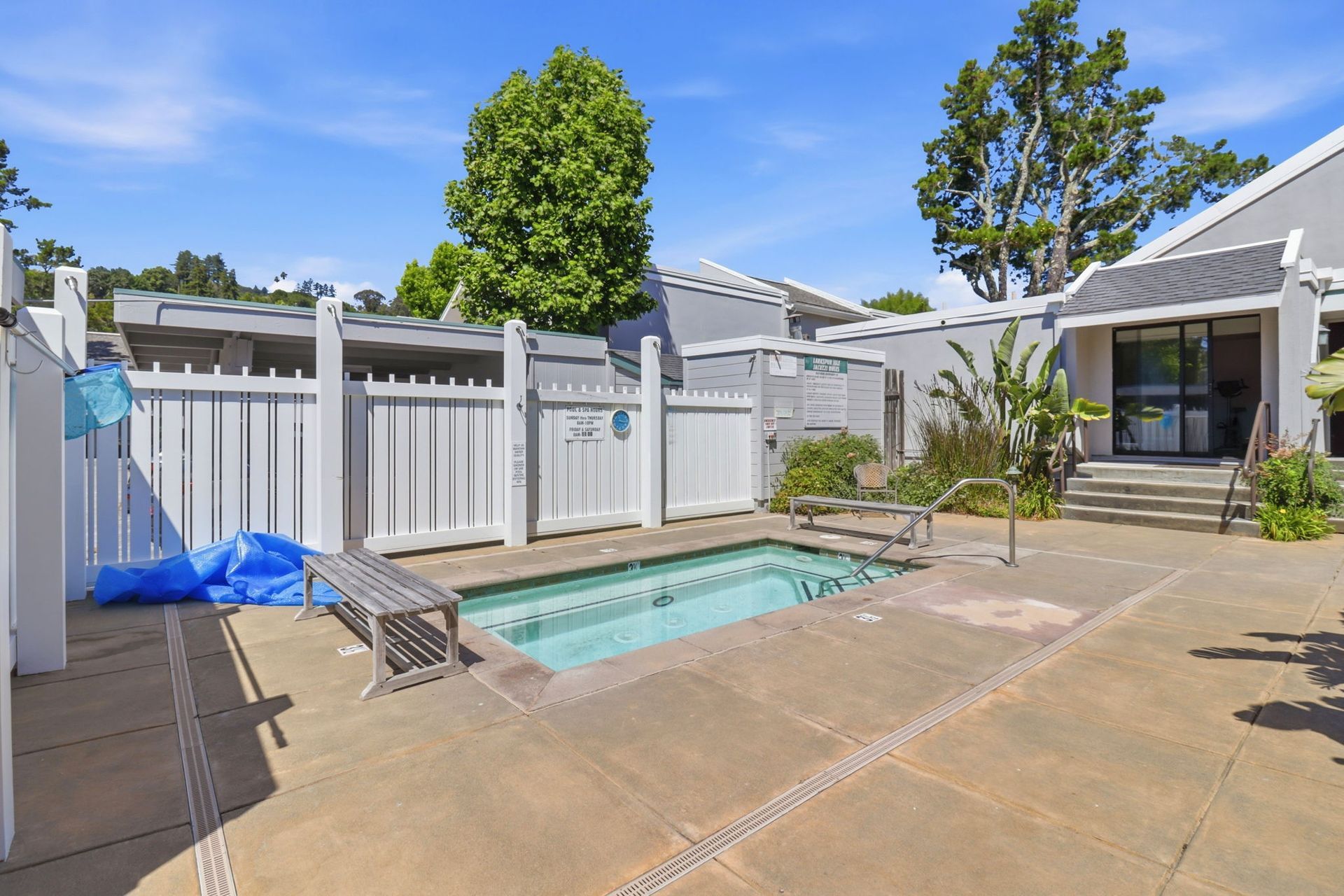 Backyard patio with a small pool, lounge chairs, white fence, and trees under a sunny sky