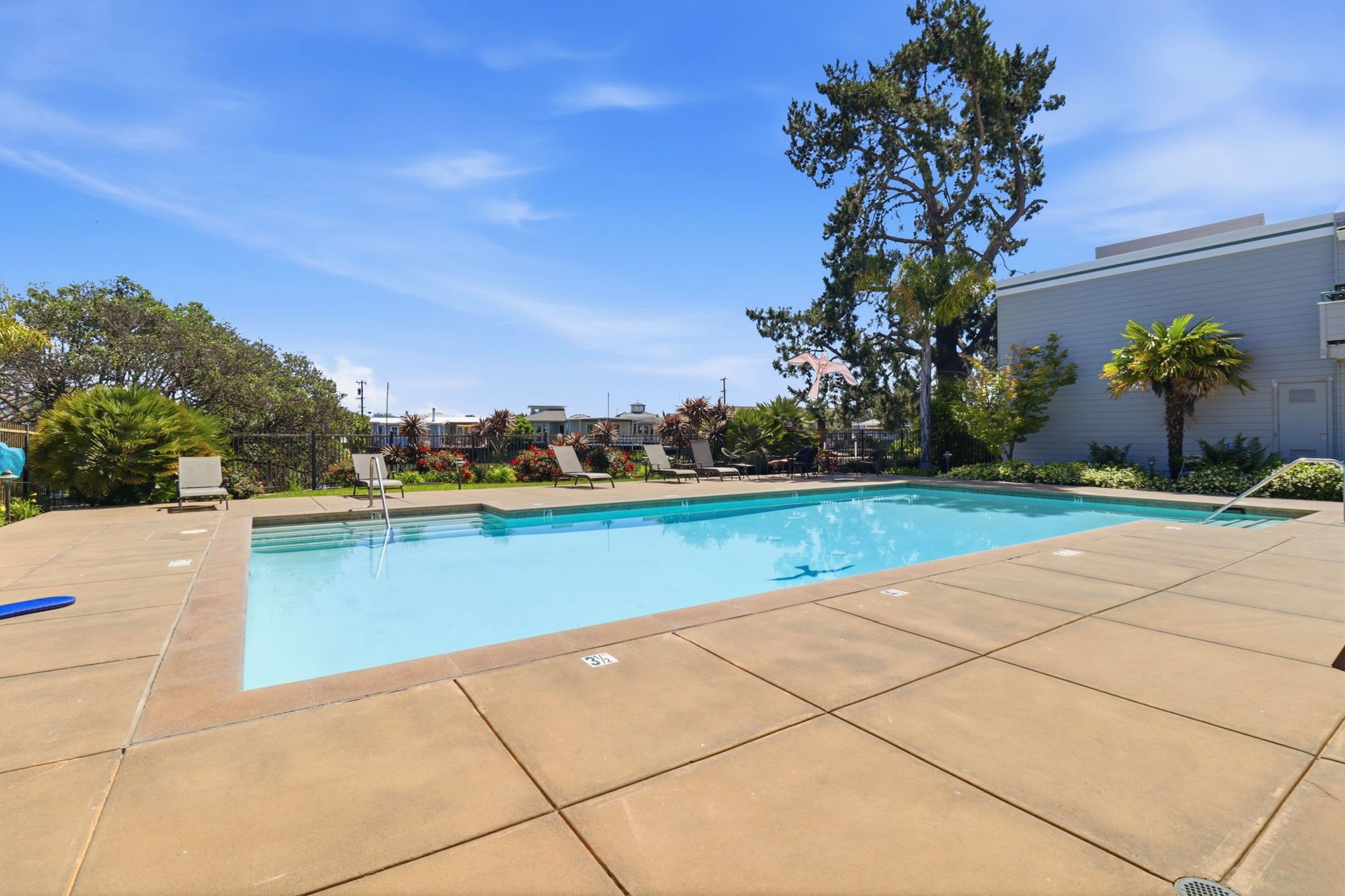 Outdoor swimming pool with clear blue water, tan deck, and trees under a bright blue sky