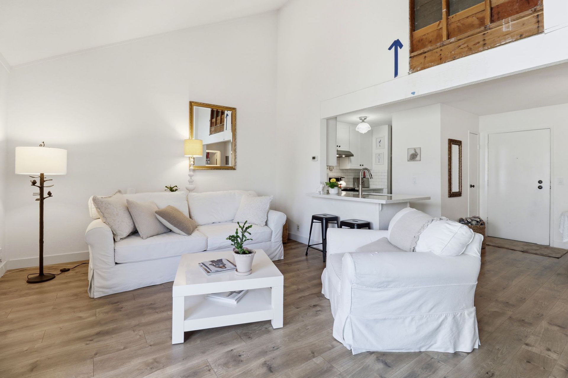 Bright living room with white sofas, coffee table, floor lamp, mirror, and high windows above a loft opening