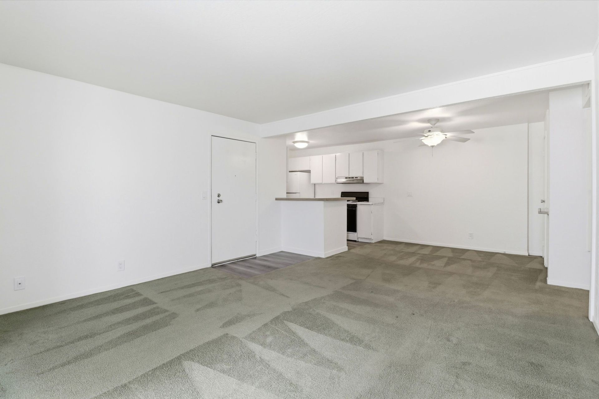 Empty living room with green carpet and open kitchen, white walls, and ceiling fan above.