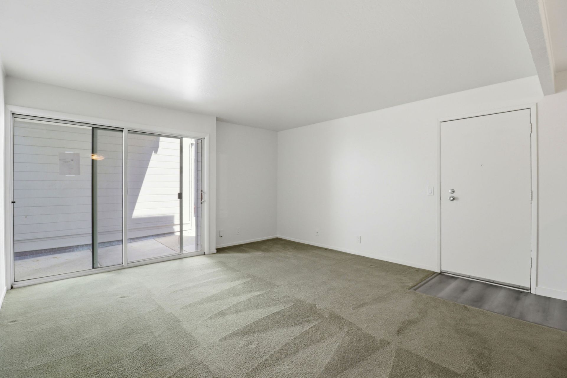 Empty carpeted living room with white walls, sliding glass door, and a closed white door