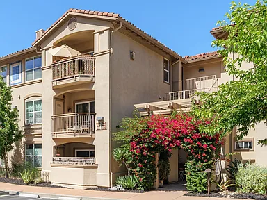 Multi-story apartment building with balconies, tan stucco, and red-flowering plants.