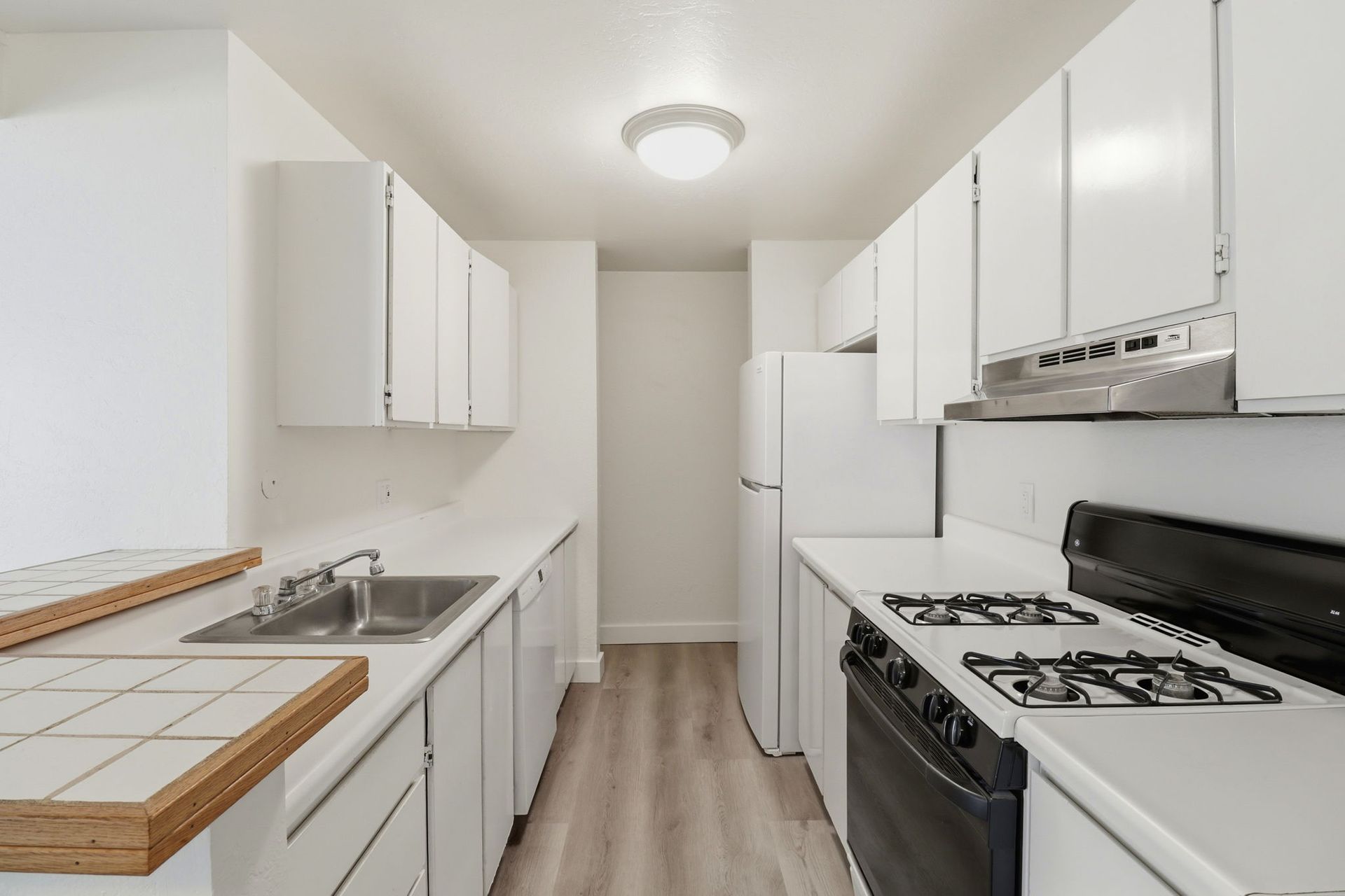 Bright white galley kitchen with tiled counters, gas stove, sink, and refrigerator