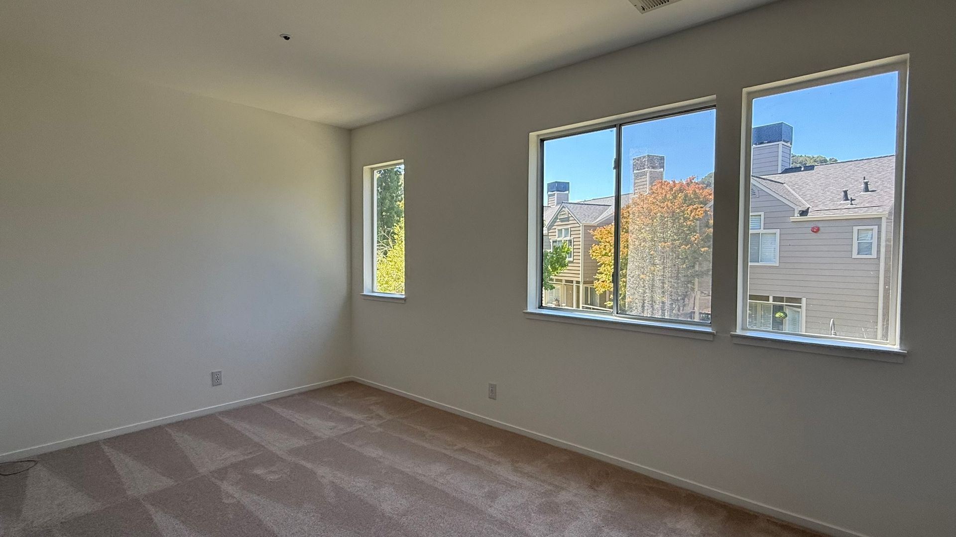 Empty beige room with carpet and three windows overlooking neighboring houses and trees