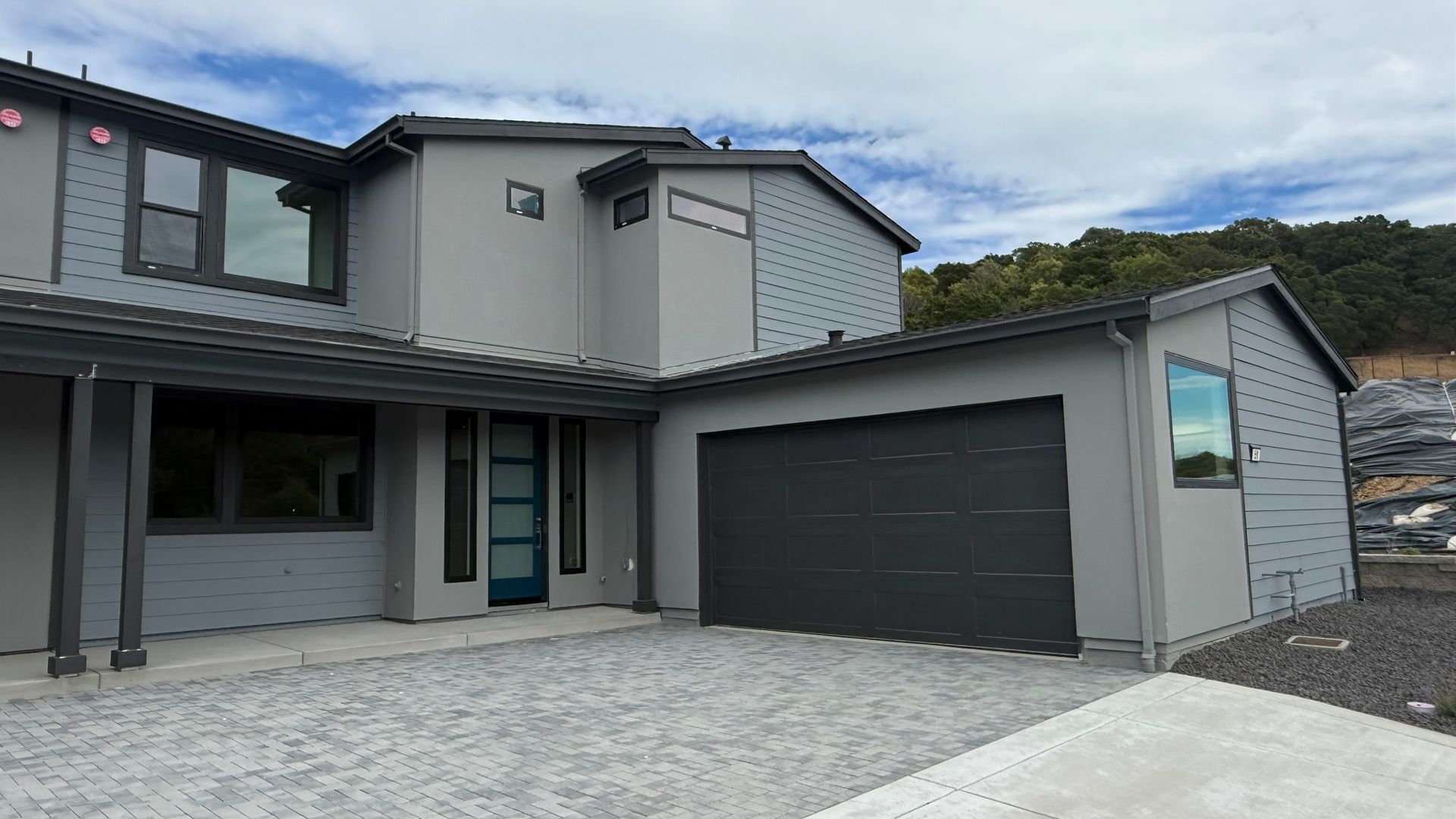 Modern gray two-story house with a dark garage door and stone driveway under a cloudy sky