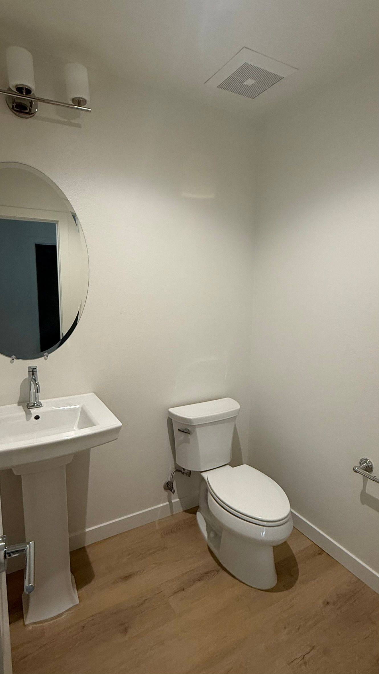 Small white bathroom with pedestal sink, toilet, mirror, and wood-look flooring.