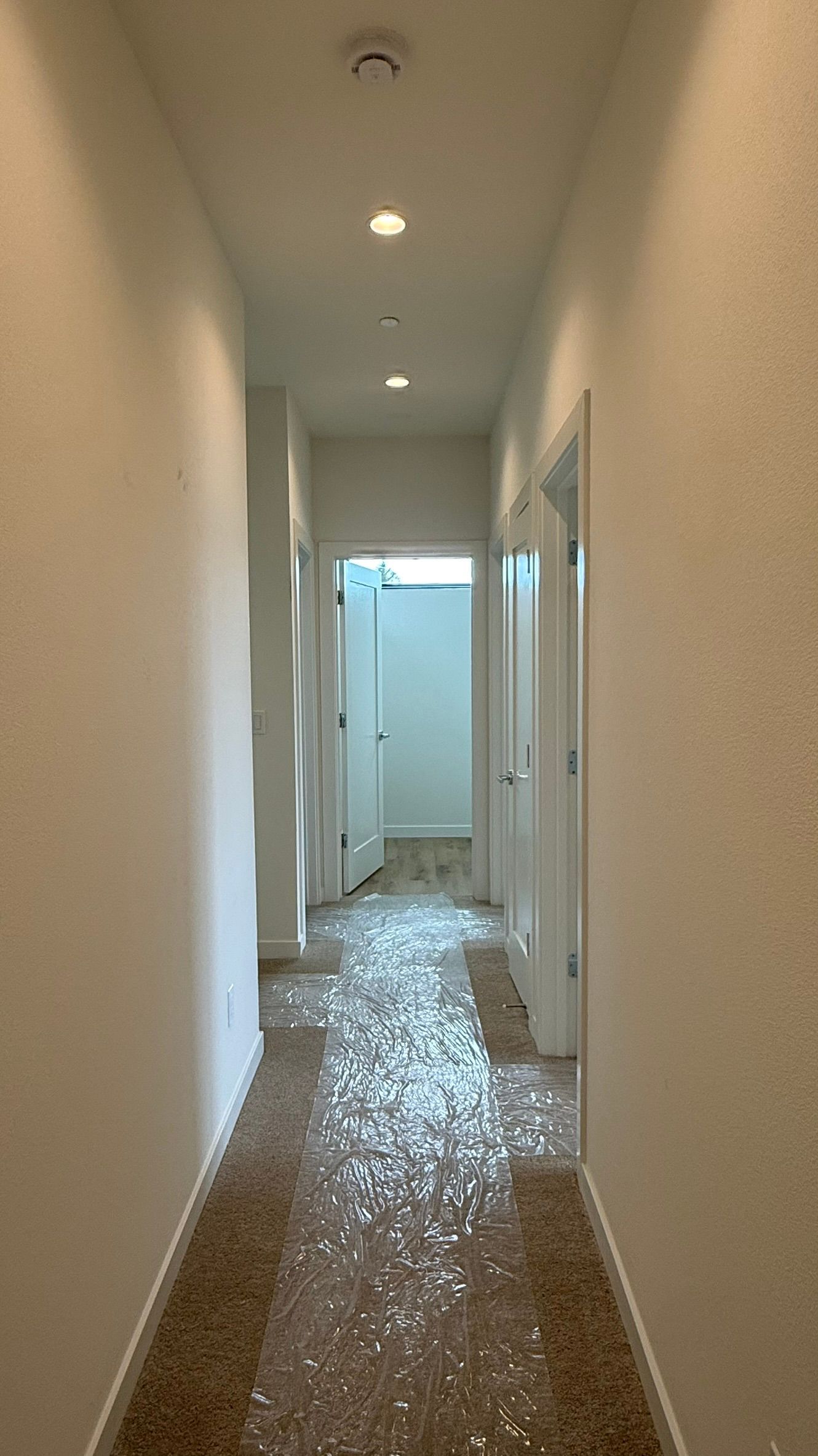 Narrow white hallway with tiled floor, ceiling lights, and doors along both sides.